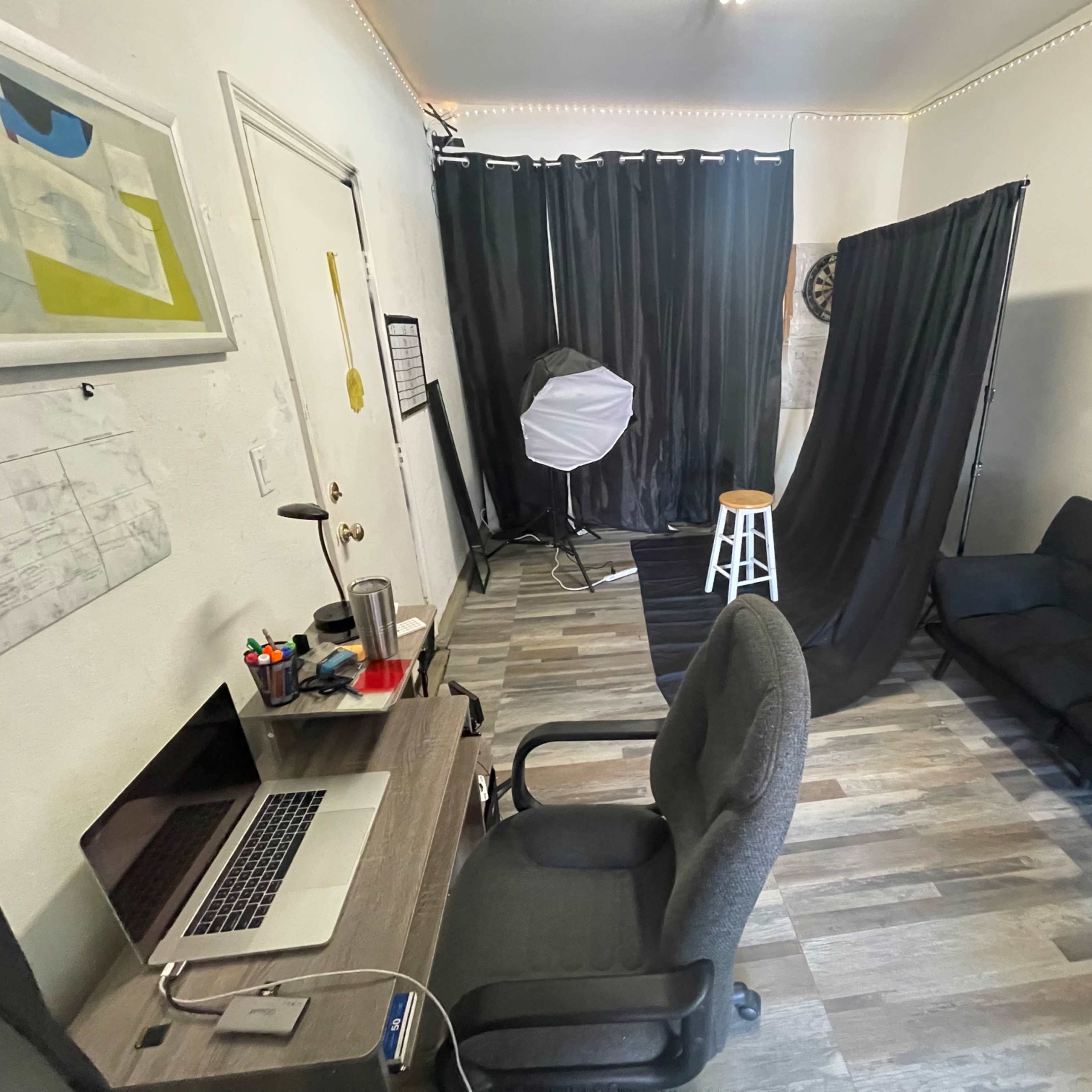 A small studio space with a desk and computer, a black backdrop and lighting equipment, and a white stool in the corner.