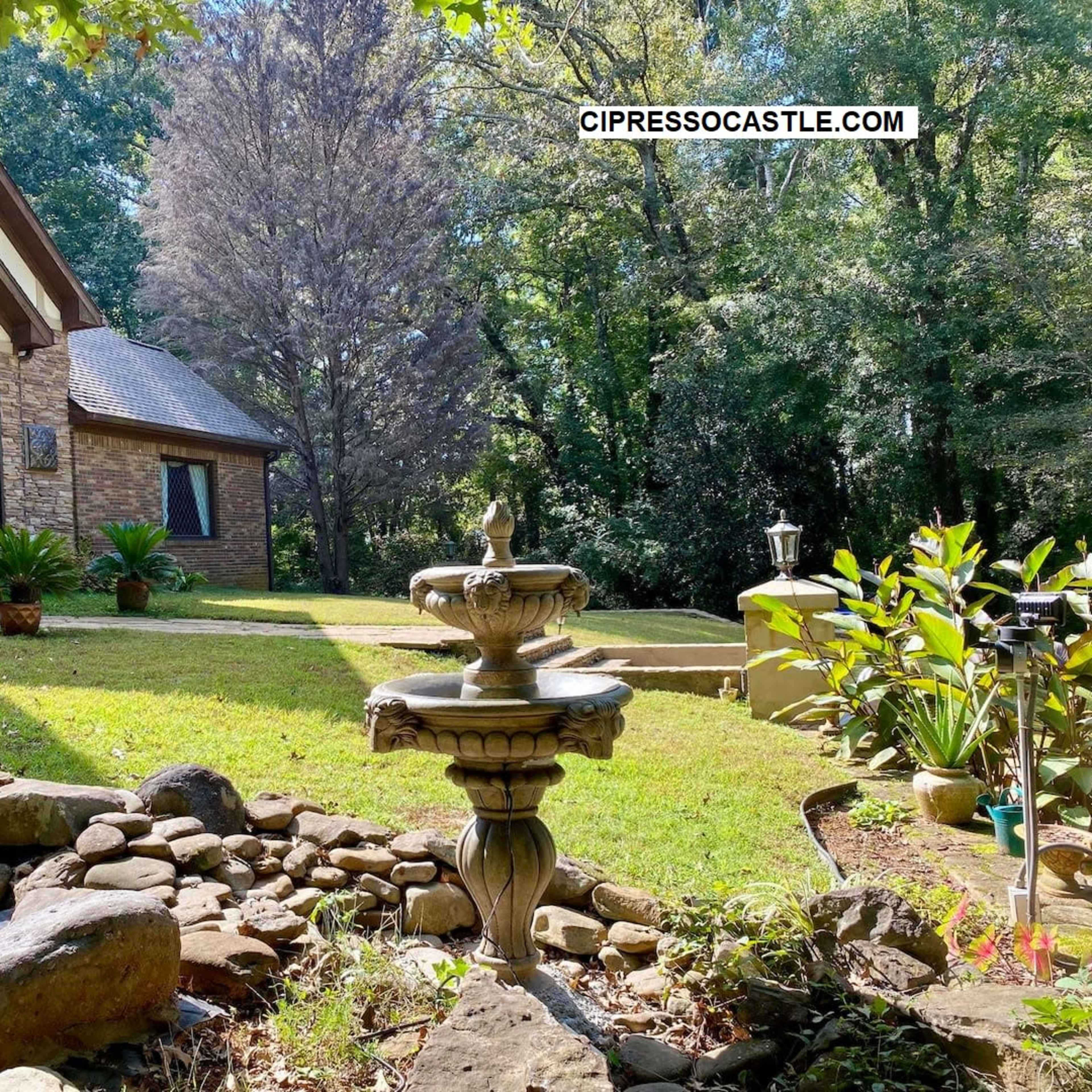 A stone fountain stands in the foreground of a landscaped yard, leading up to a house surrounded by trees.