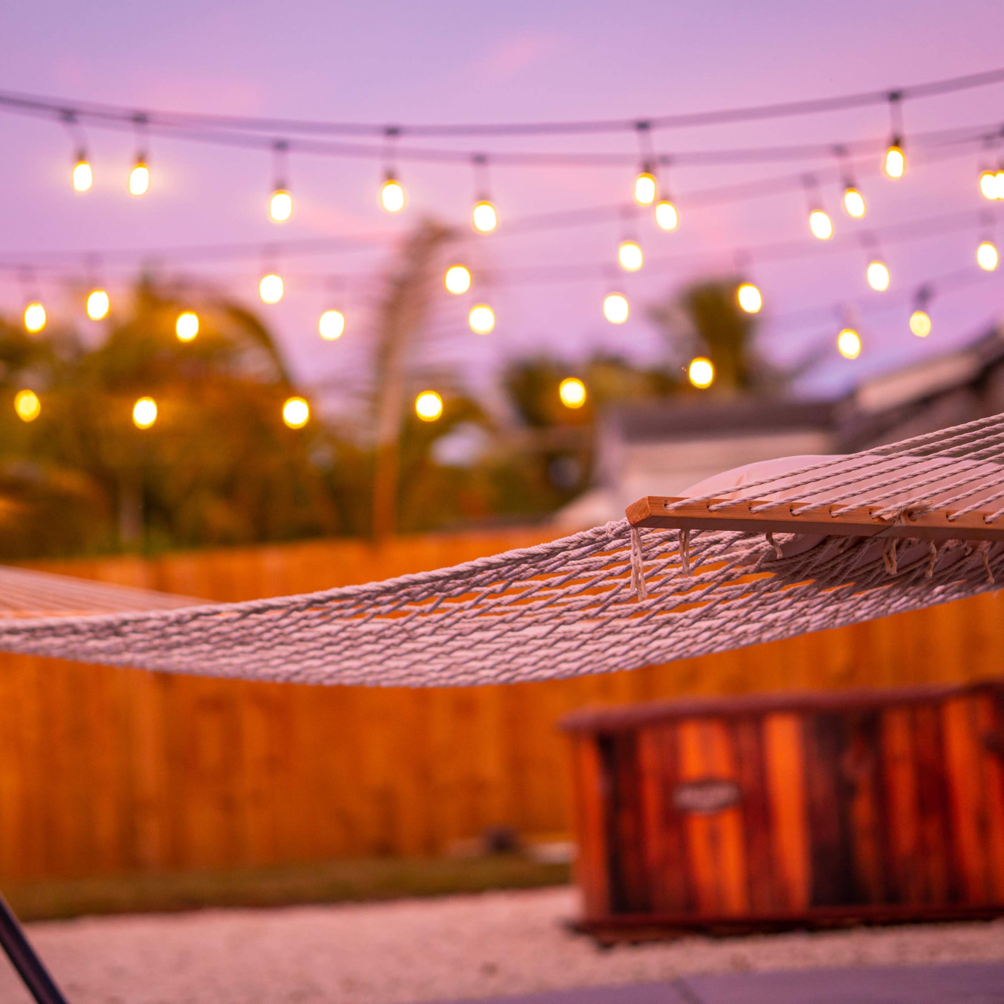 A hammock is suspended near a wooden hot tub, with string lights illuminated in the background against a twilight sky.