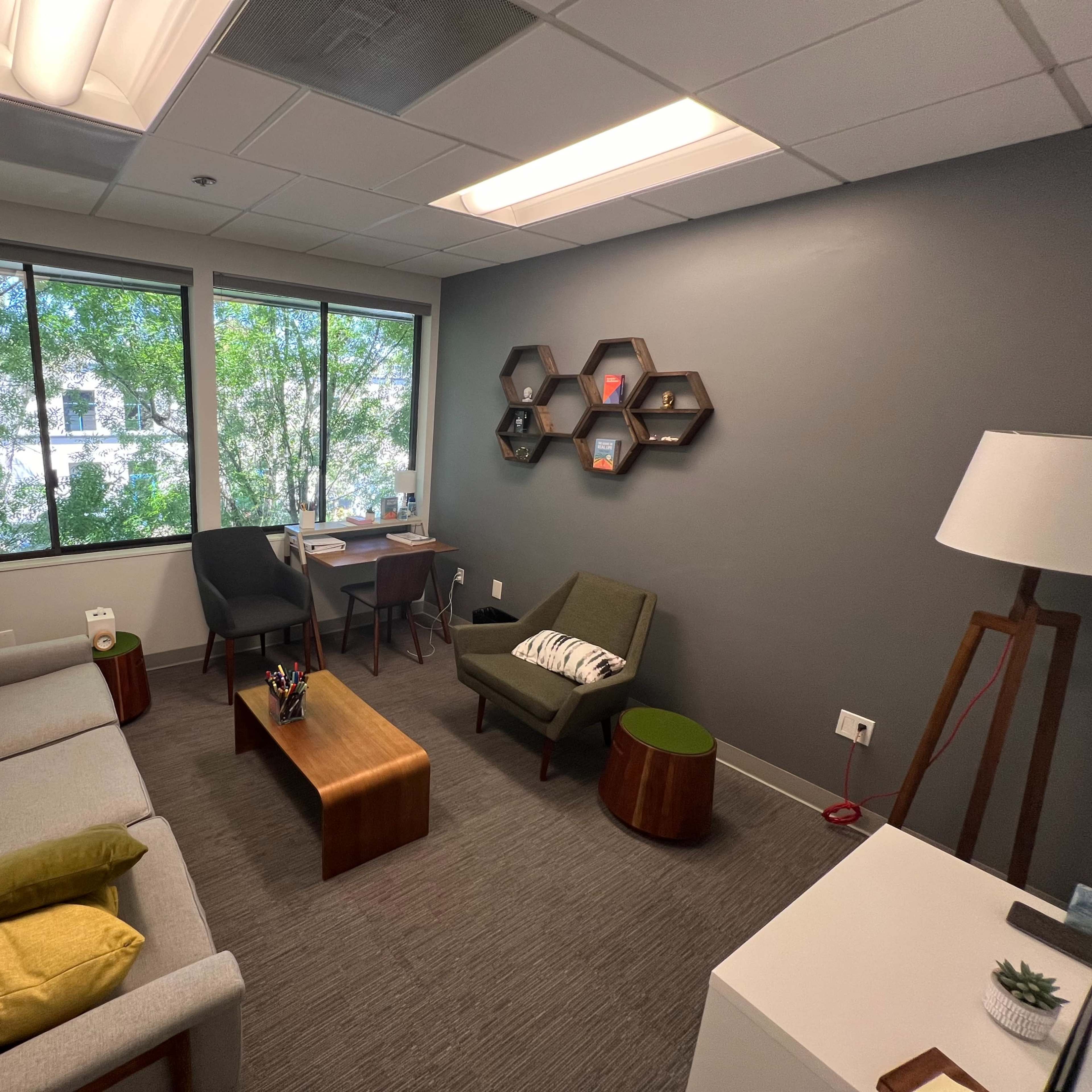 A cozy office space features a grey sofa, two chairs, a wooden coffee table, and a desk, with large windows allowing natural light and greenery to enter.