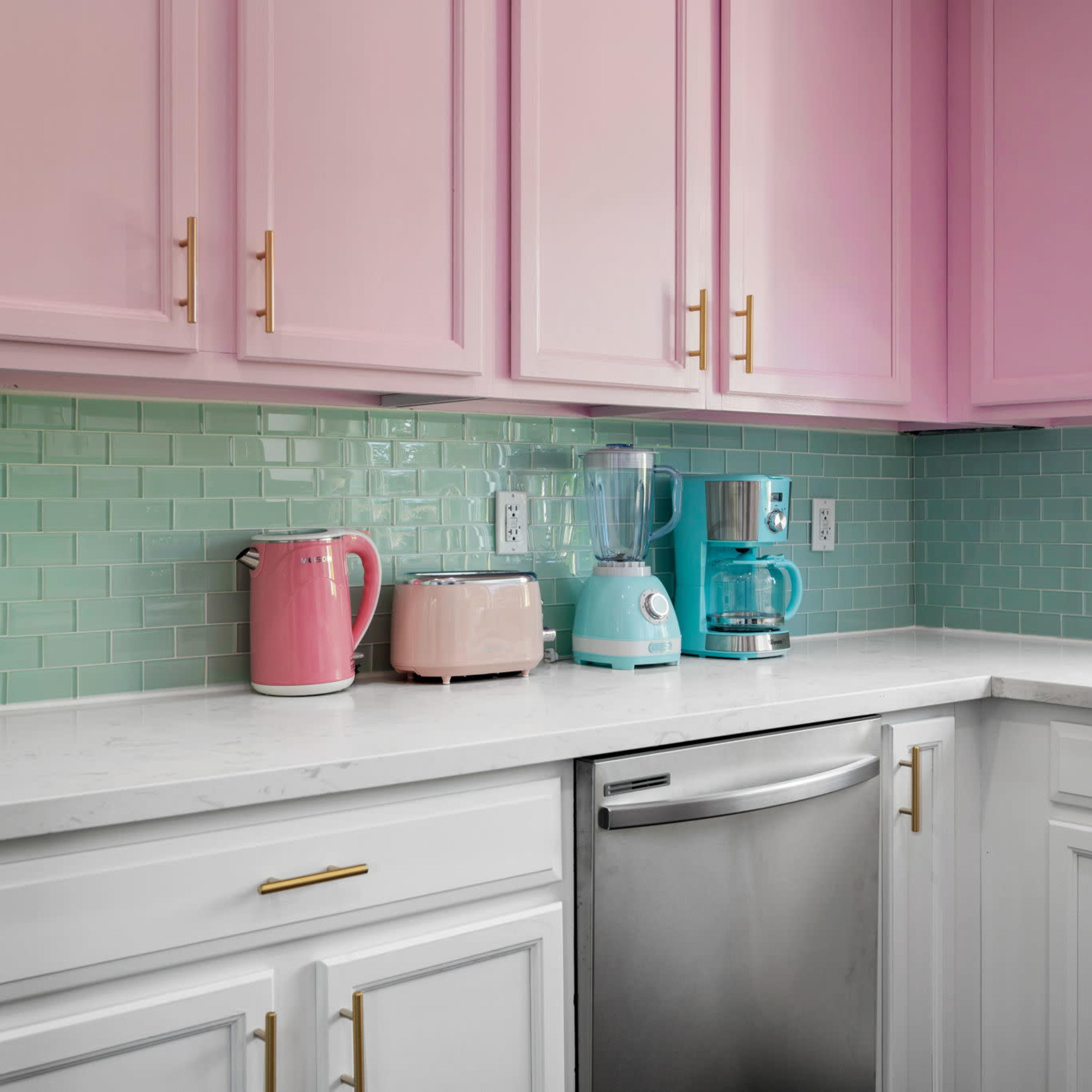 A kitchen with pink upper cabinets, a white lower cabinet, and mint green tile backsplash, featuring a pink kettle, a cream toaster, a blue coffee maker, and a blender.