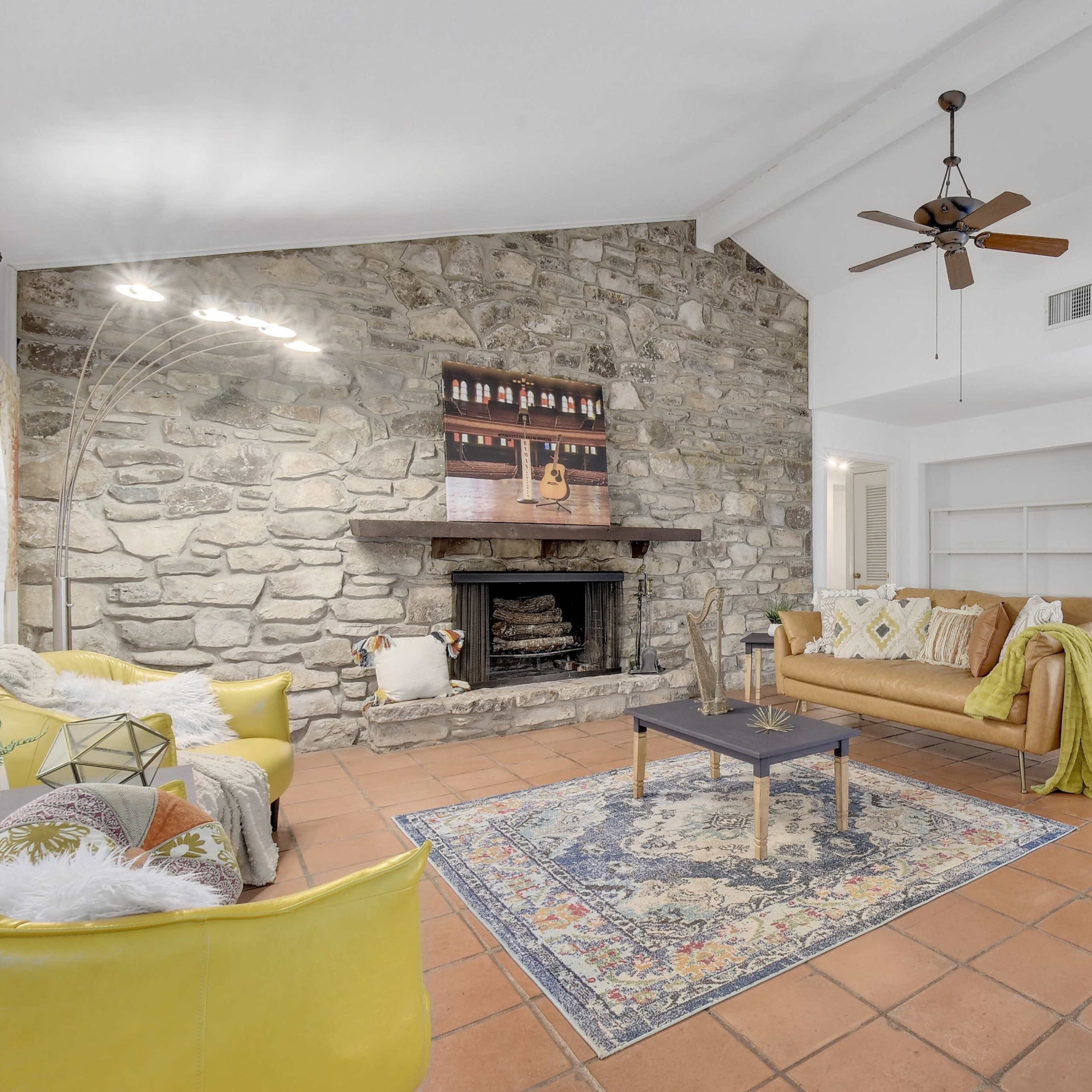 The living room features a stone fireplace, a wooden sofa with decorative pillows, and a patterned rug on terracotta tile flooring.