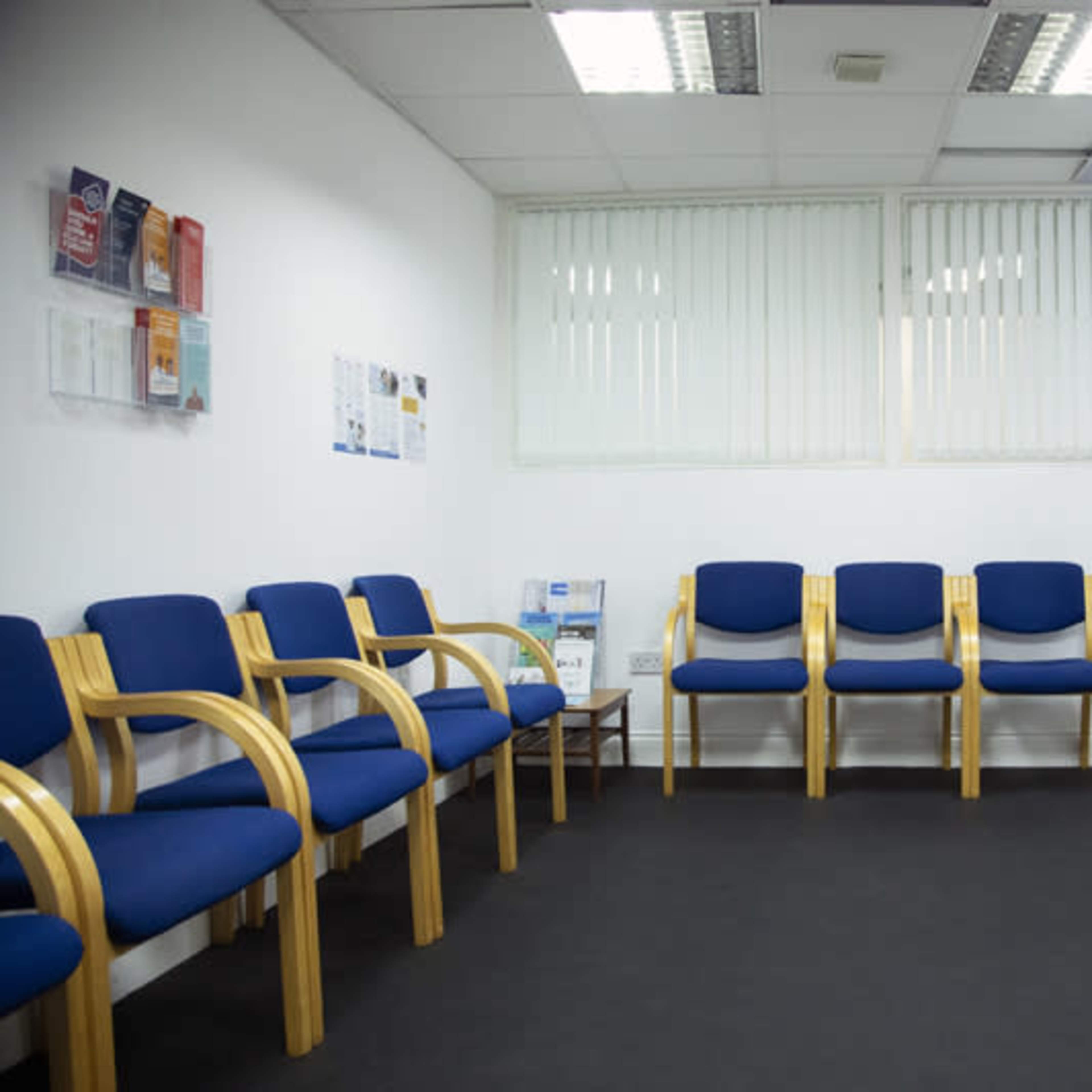 A waiting room with several blue chairs arranged along the wall and a small table with pamphlets in a well-lit space.