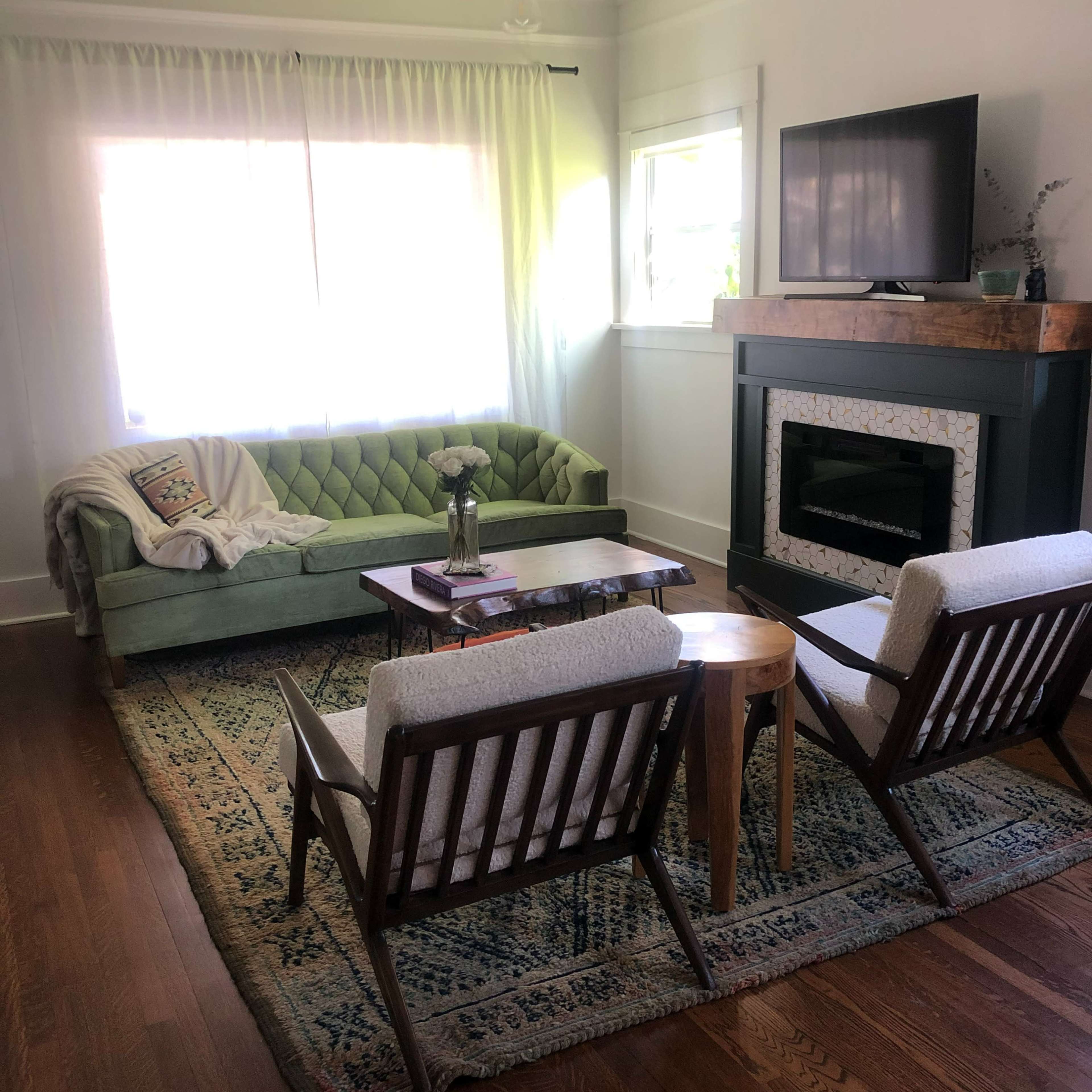 A cozy living room features a green tufted sofa, two wooden chairs, a wooden coffee table, and a fireplace with a light-colored mantel.