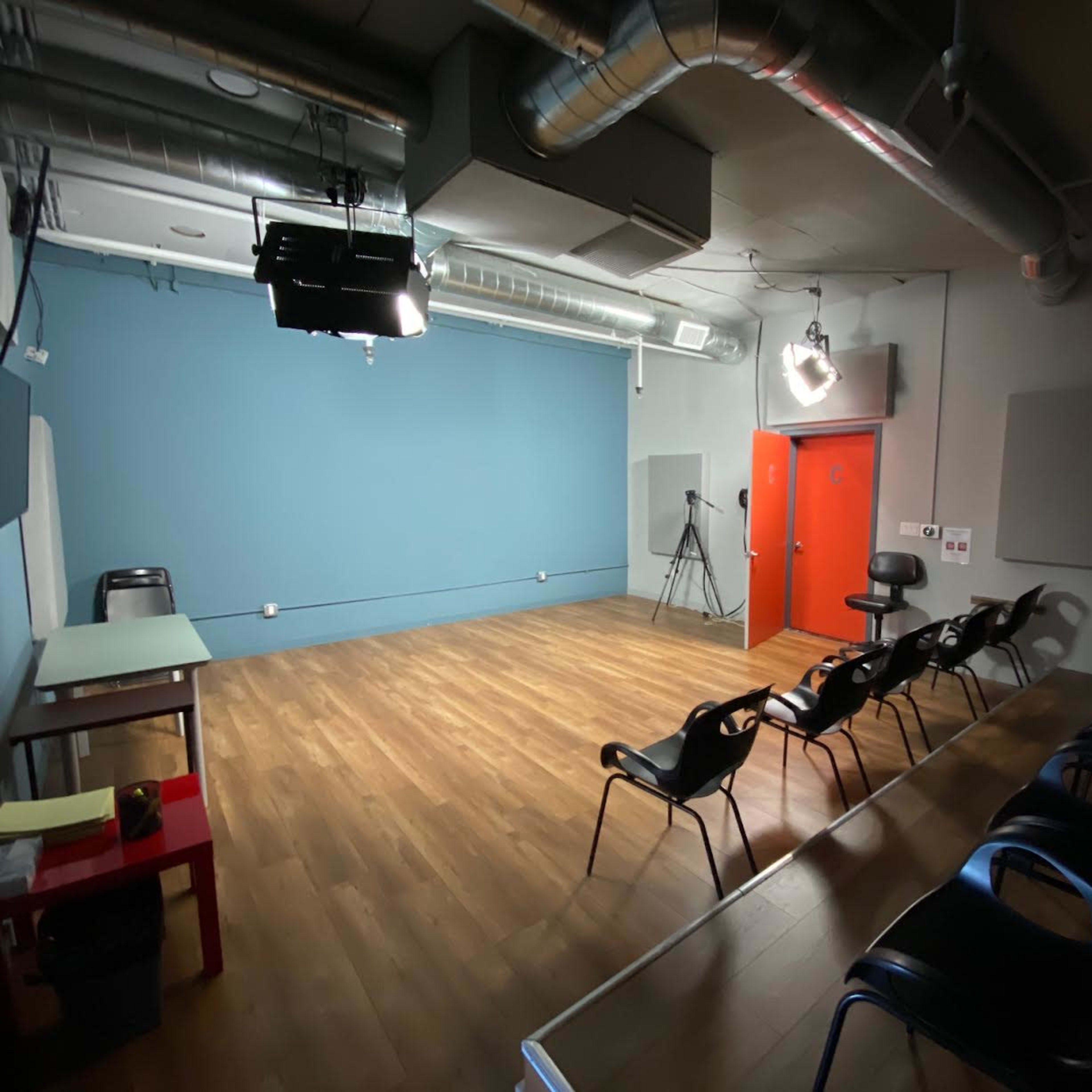 An empty rehearsal studio with a blue wall, wooden flooring, a chair and table, and several black chairs arranged in a row.