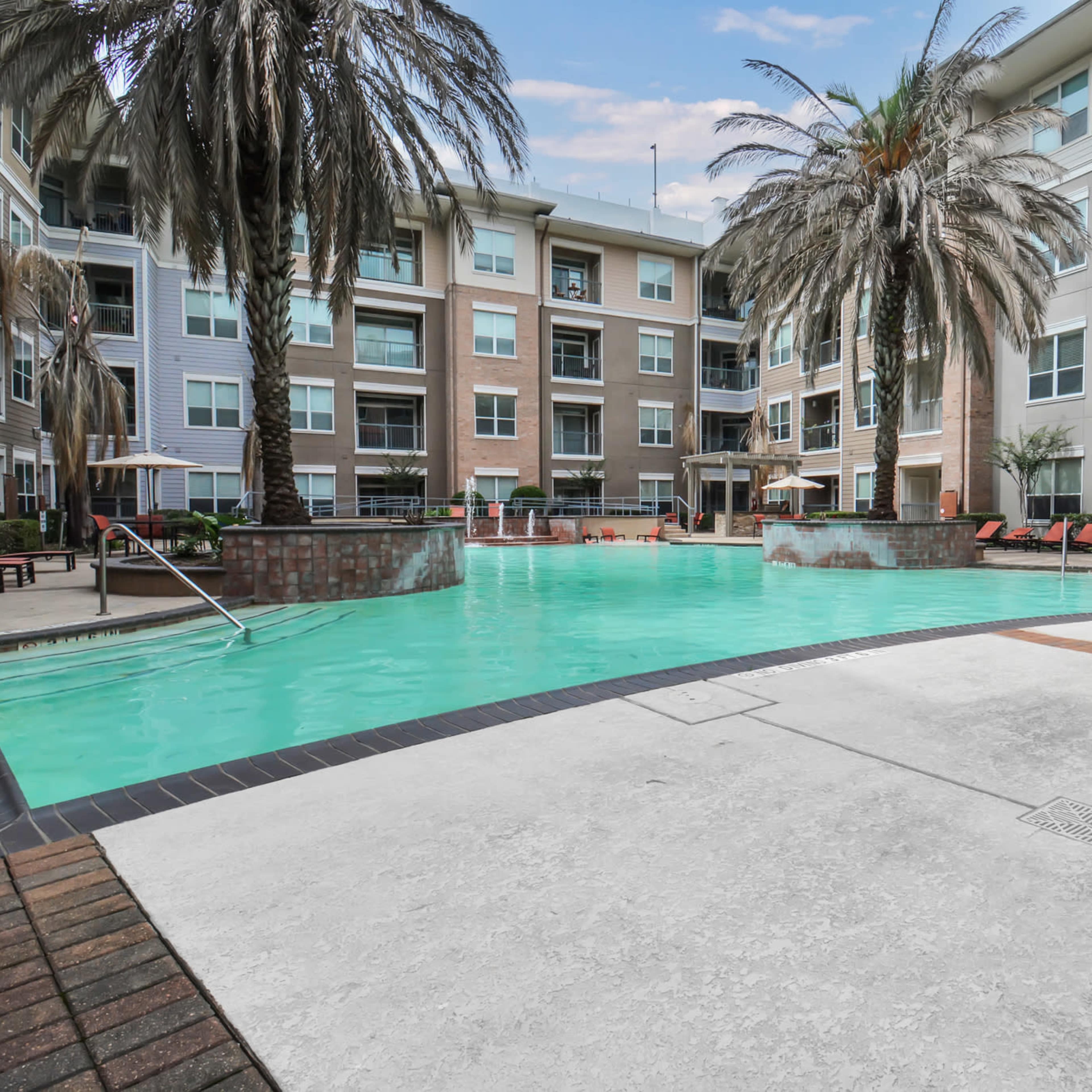 A landscaped pool area surrounded by palm trees and residential buildings, with lounge chairs positioned along the water's edge.