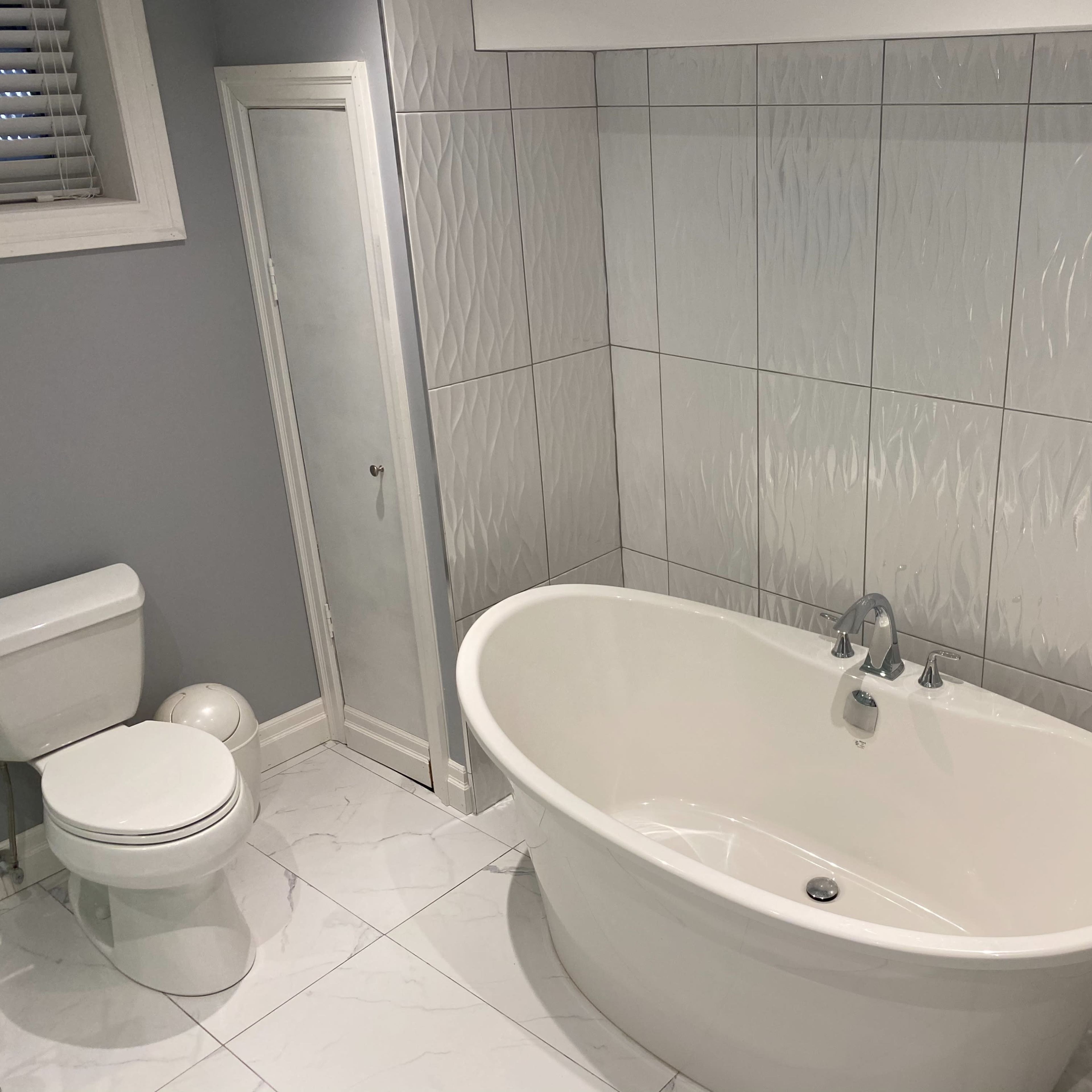 A bathroom with a freestanding white tub, a toilet, and textured wall tiles in a light-colored setting.