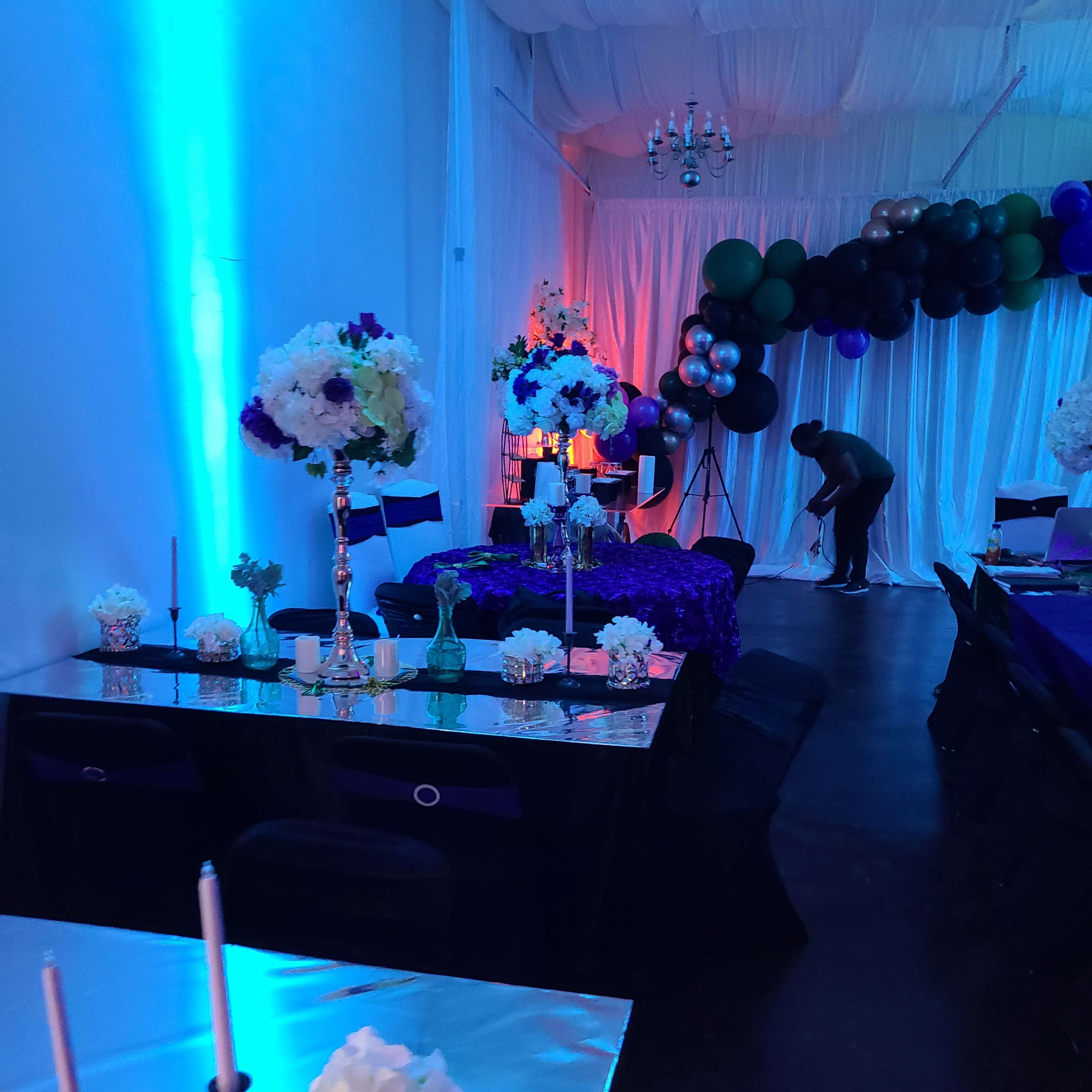 A decorated indoor event space features tables with floral centerpieces, candles, and colorful lighting along with a backdrop of balloons.