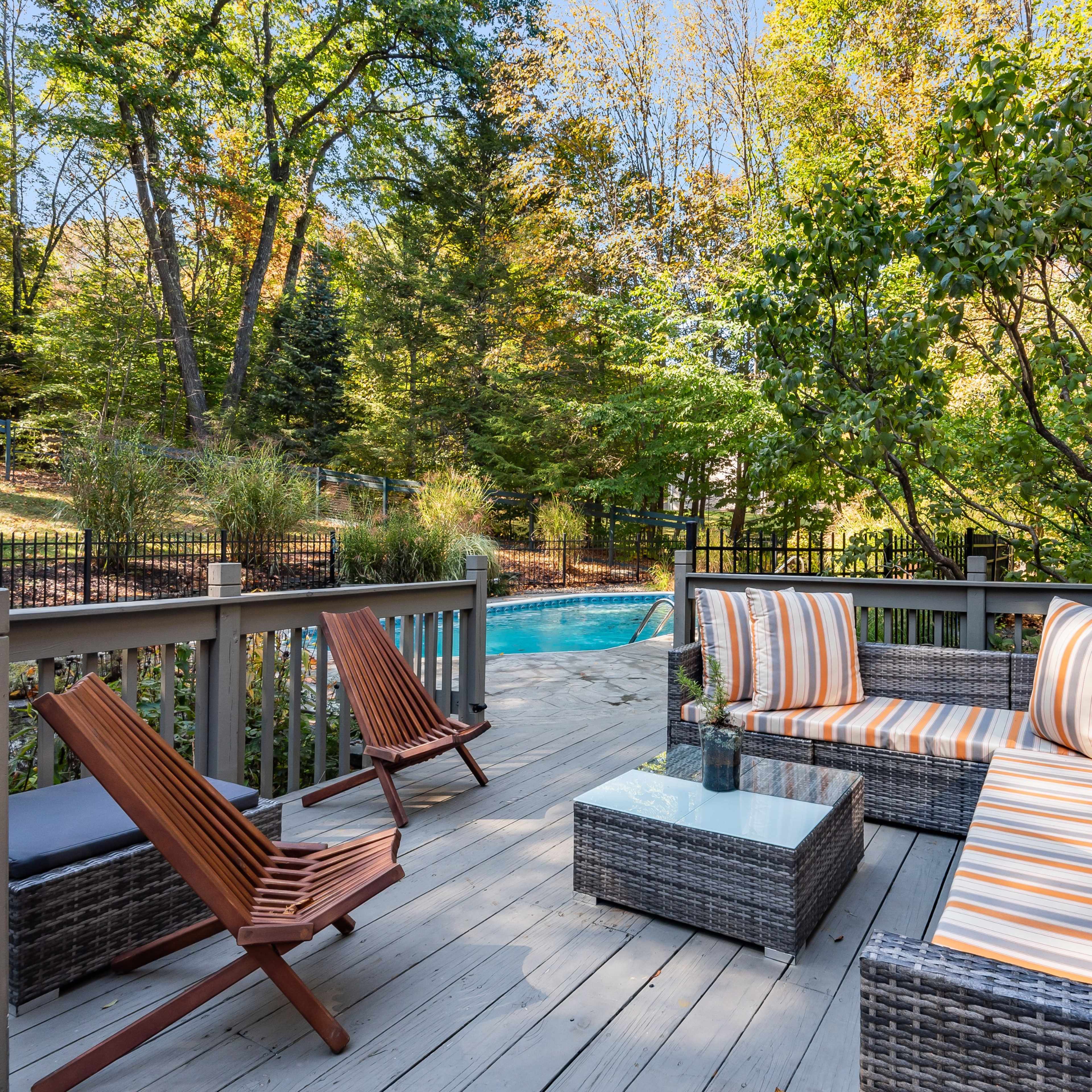 A patio area features striped cushions on wicker furniture, with a glass table and two wooden lounge chairs, adjacent to a pool surrounded by greenery.