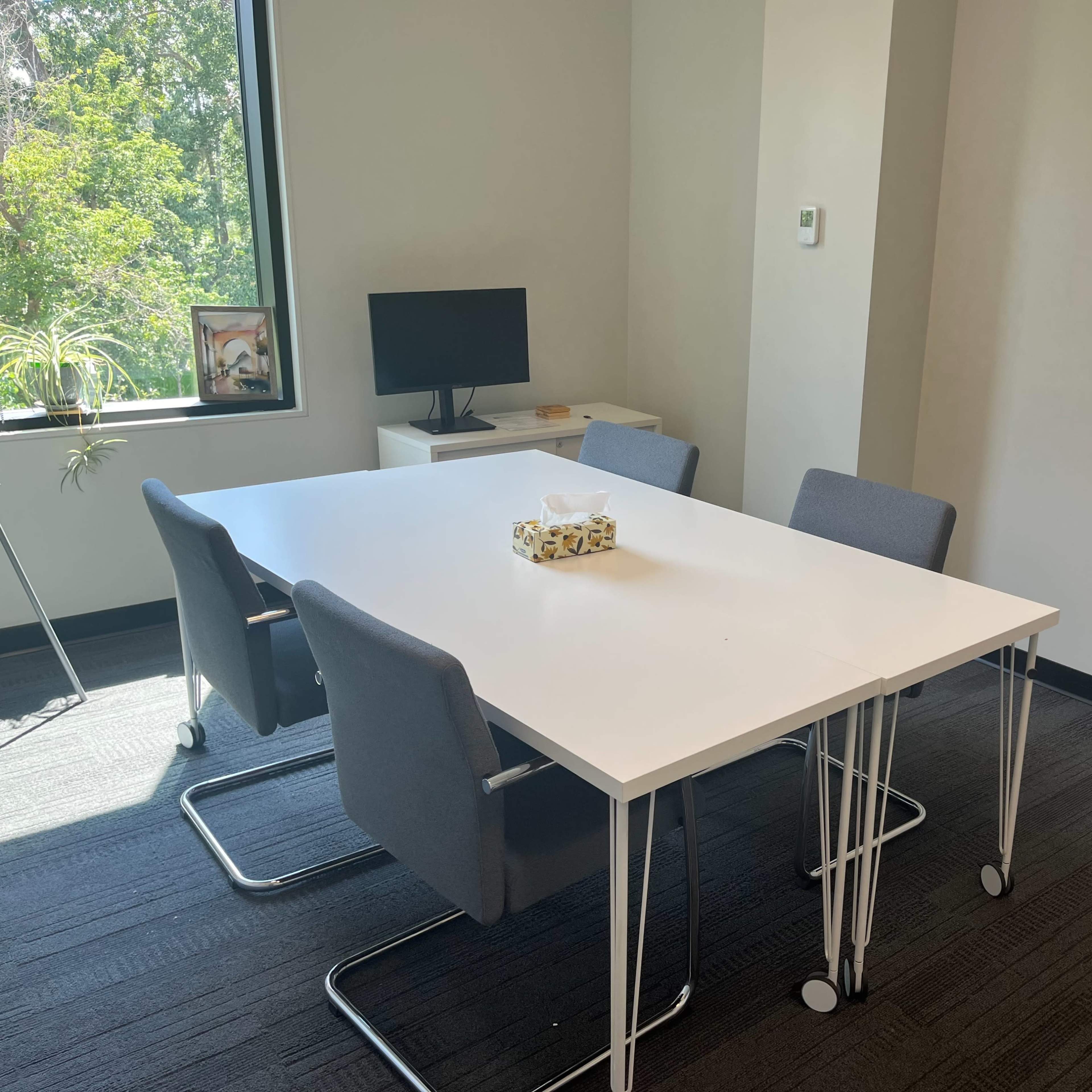 A modern meeting room featuring a white table surrounded by four gray chairs, a television on a side table, and a window allowing natural light.