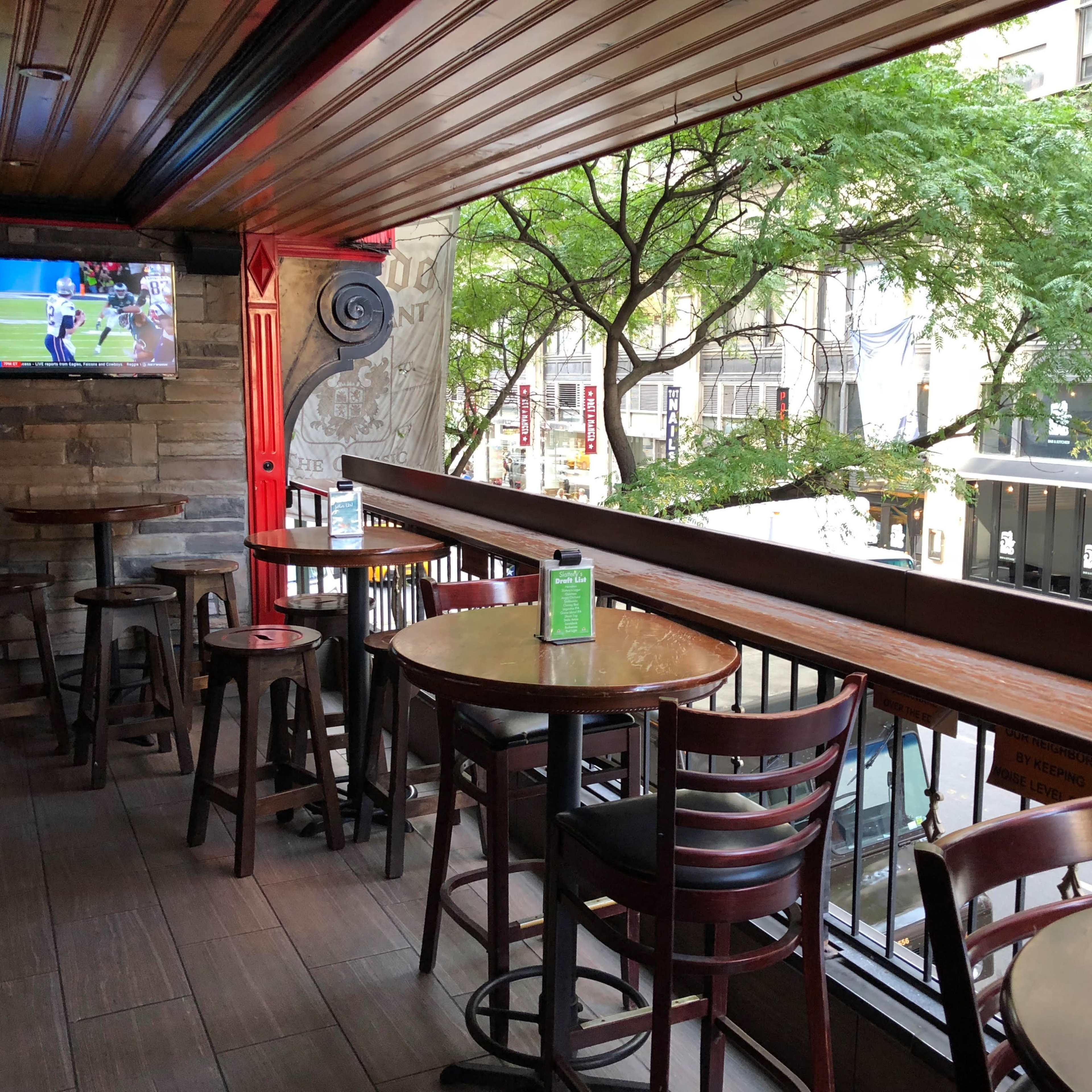 A rooftop outdoor seating area of a bar with wooden tables and chairs, a small TV mounted on the wall, and views of city buildings and trees.