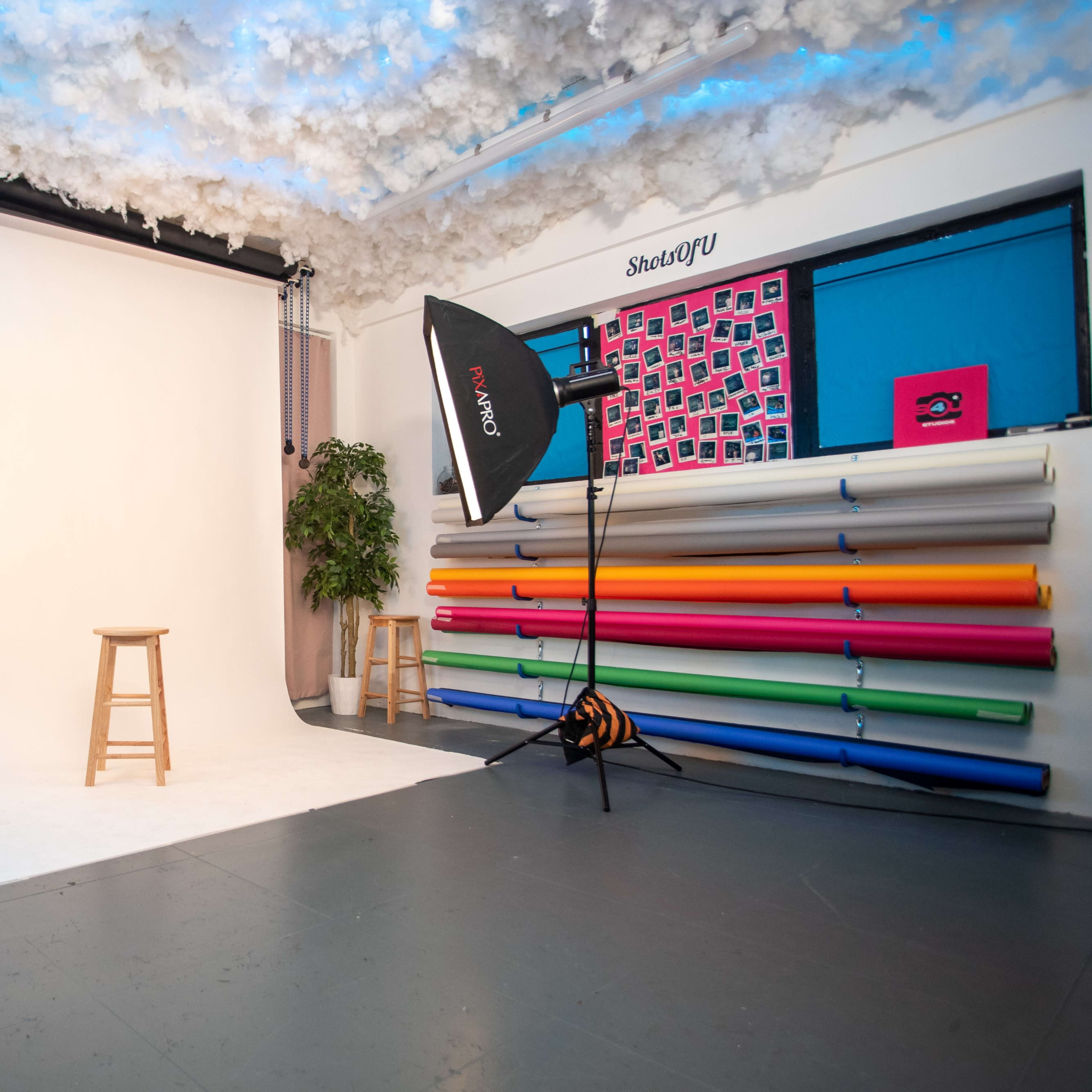 A photography studio with a blank backdrop, two softbox lights, a stool, and a variety of colored backdrop rolls.