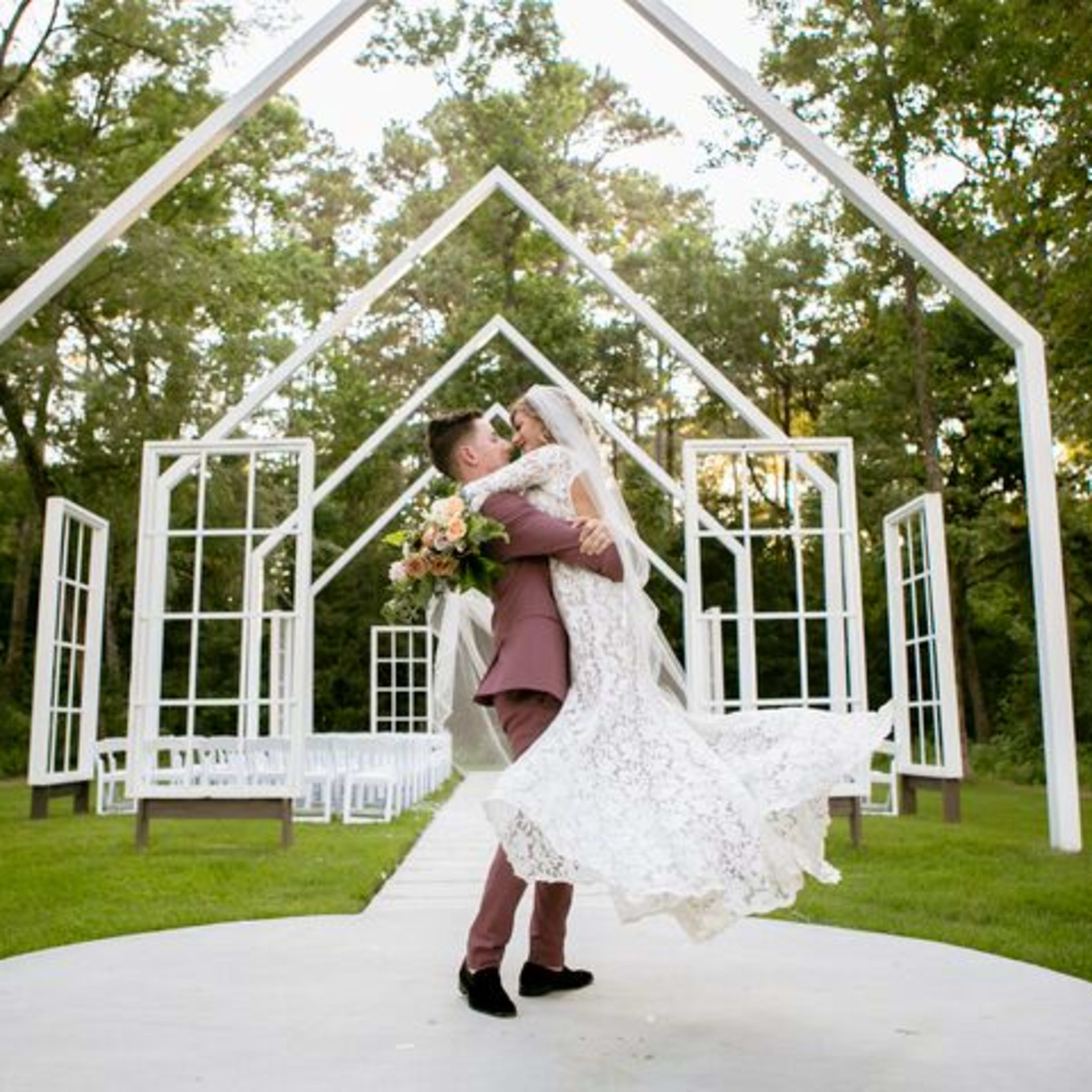 A groom lifts his bride in front of a modern wedding altar framed by trees.