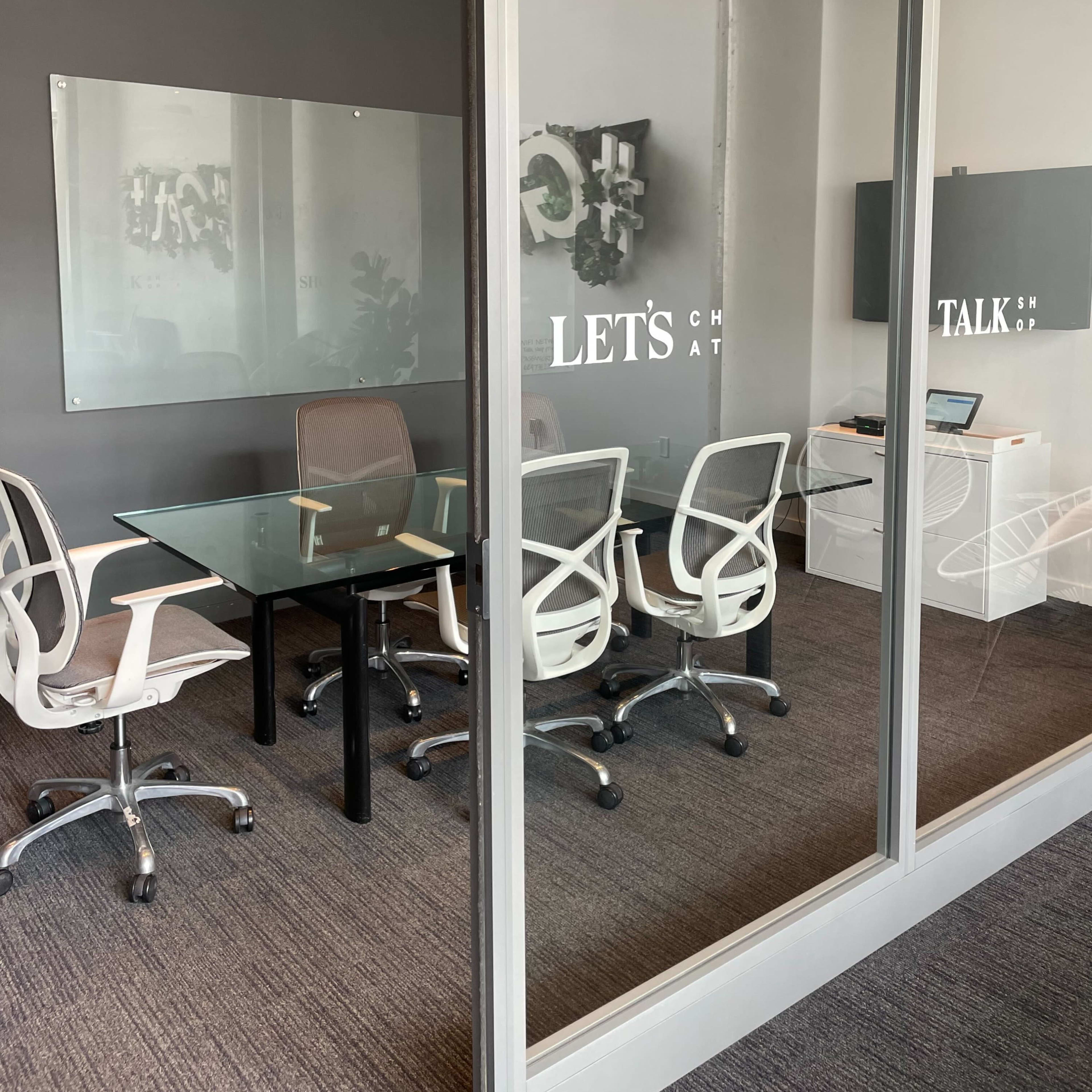 A modern conference room with a glass wall, featuring a rectangular glass table surrounded by white chairs and a television on the wall.