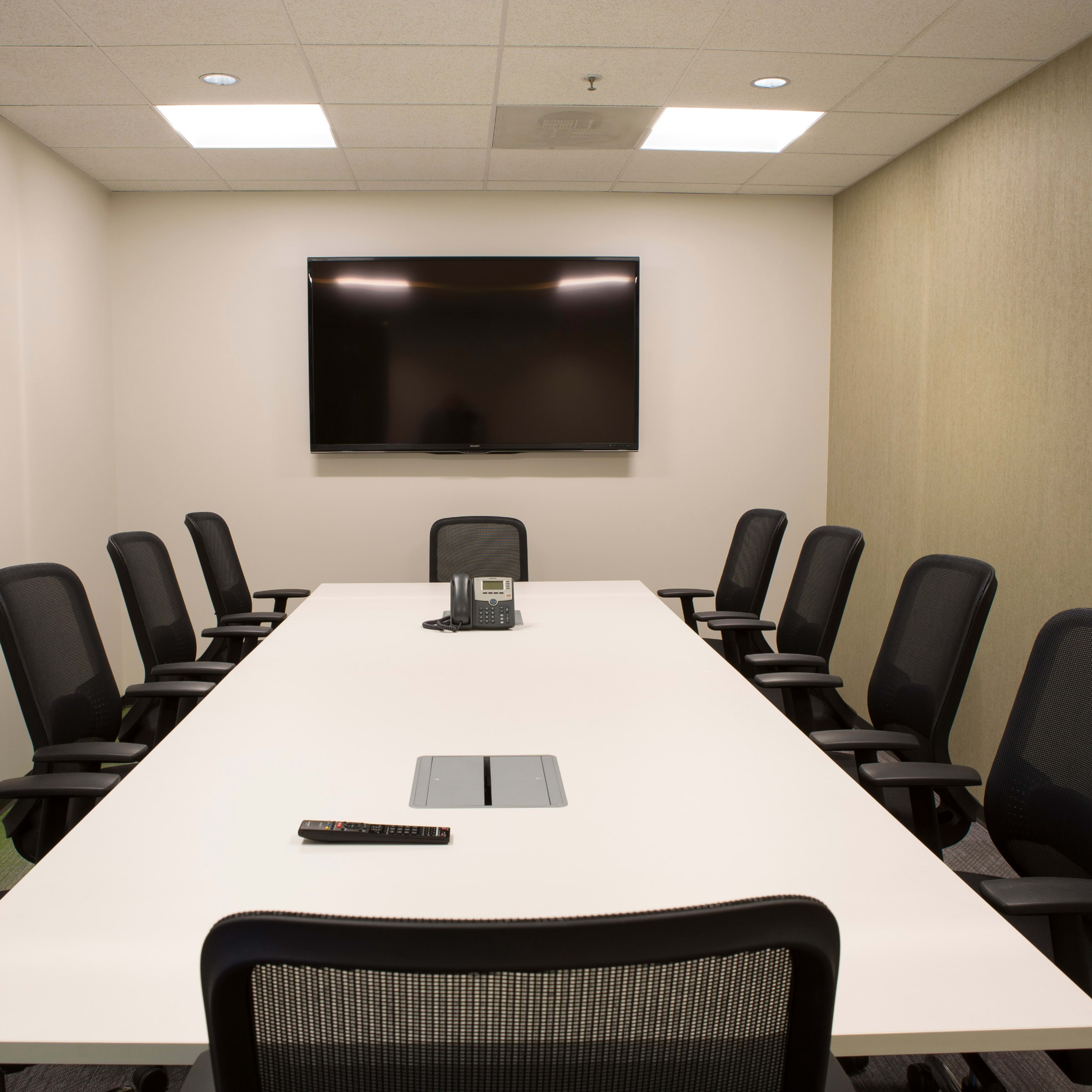 A modern conference room features a long white table surrounded by eight black mesh chairs, with a flat-screen TV mounted on the wall and a phone on the table.