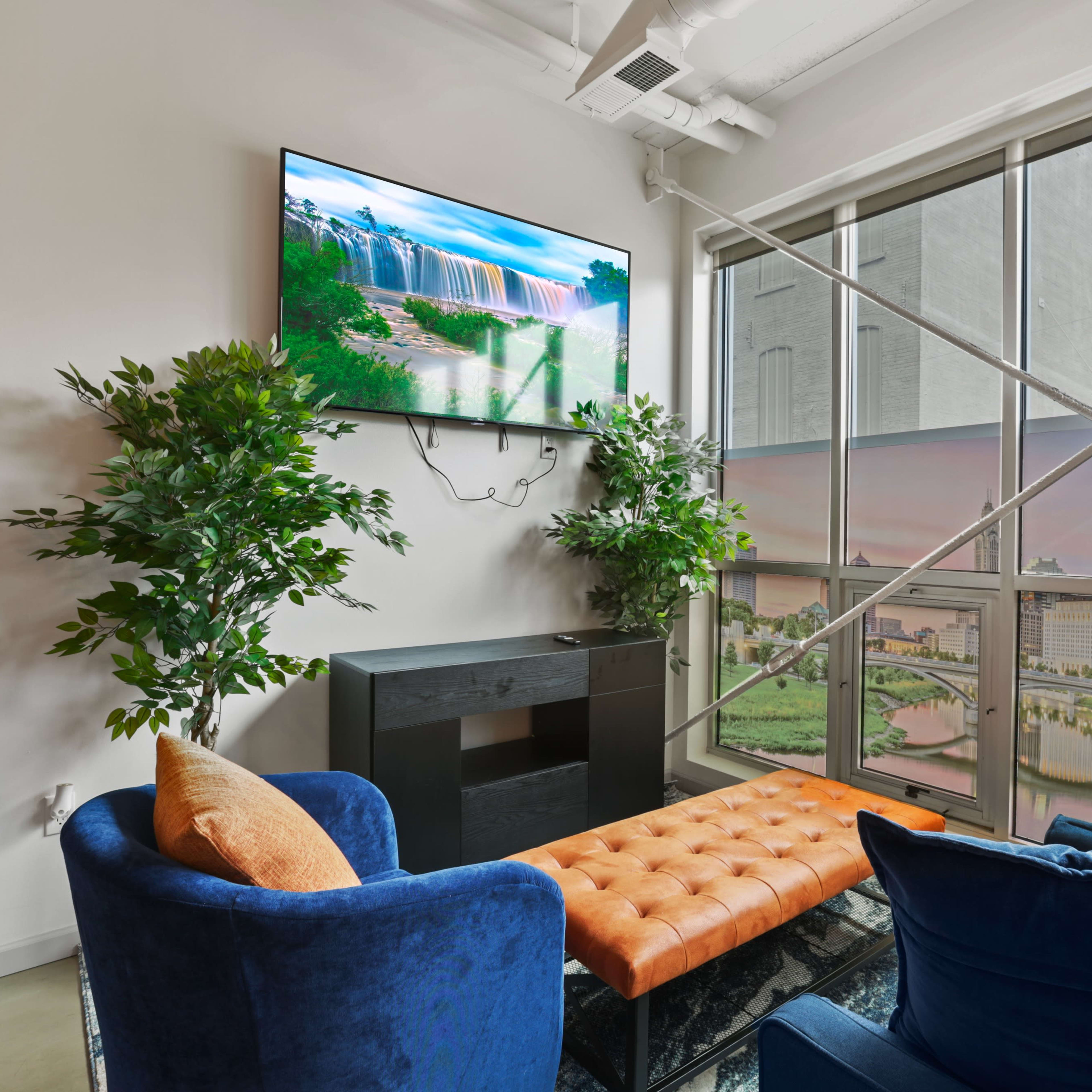 A modern living space with a large TV mounted on the wall, a black media console, two blue chairs, and a tufted orange ottoman, all positioned near a large window with a cityscape view.