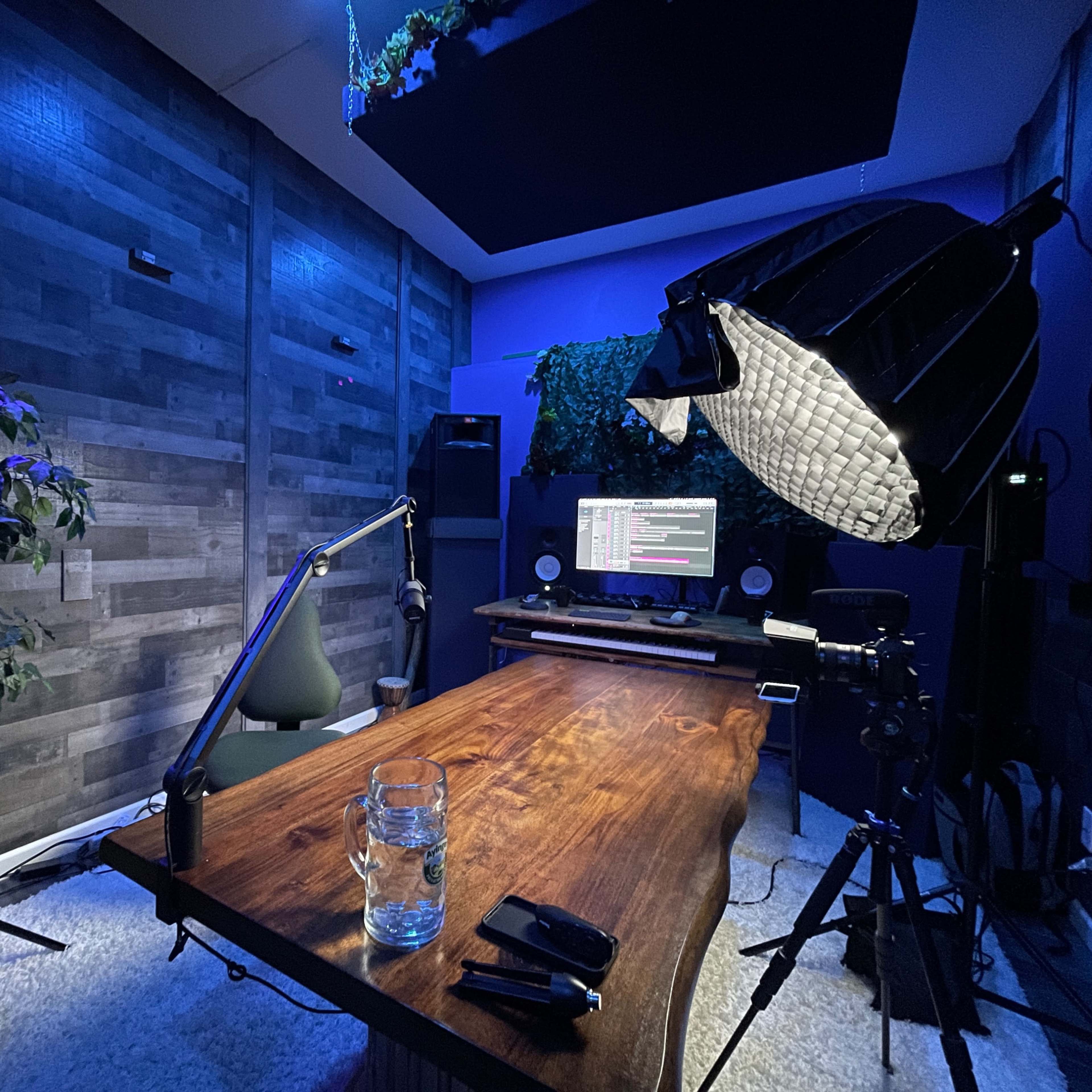 A modern recording studio with a wooden table, a computer monitor, and a large softbox light, surrounded by plants and acoustic paneling.