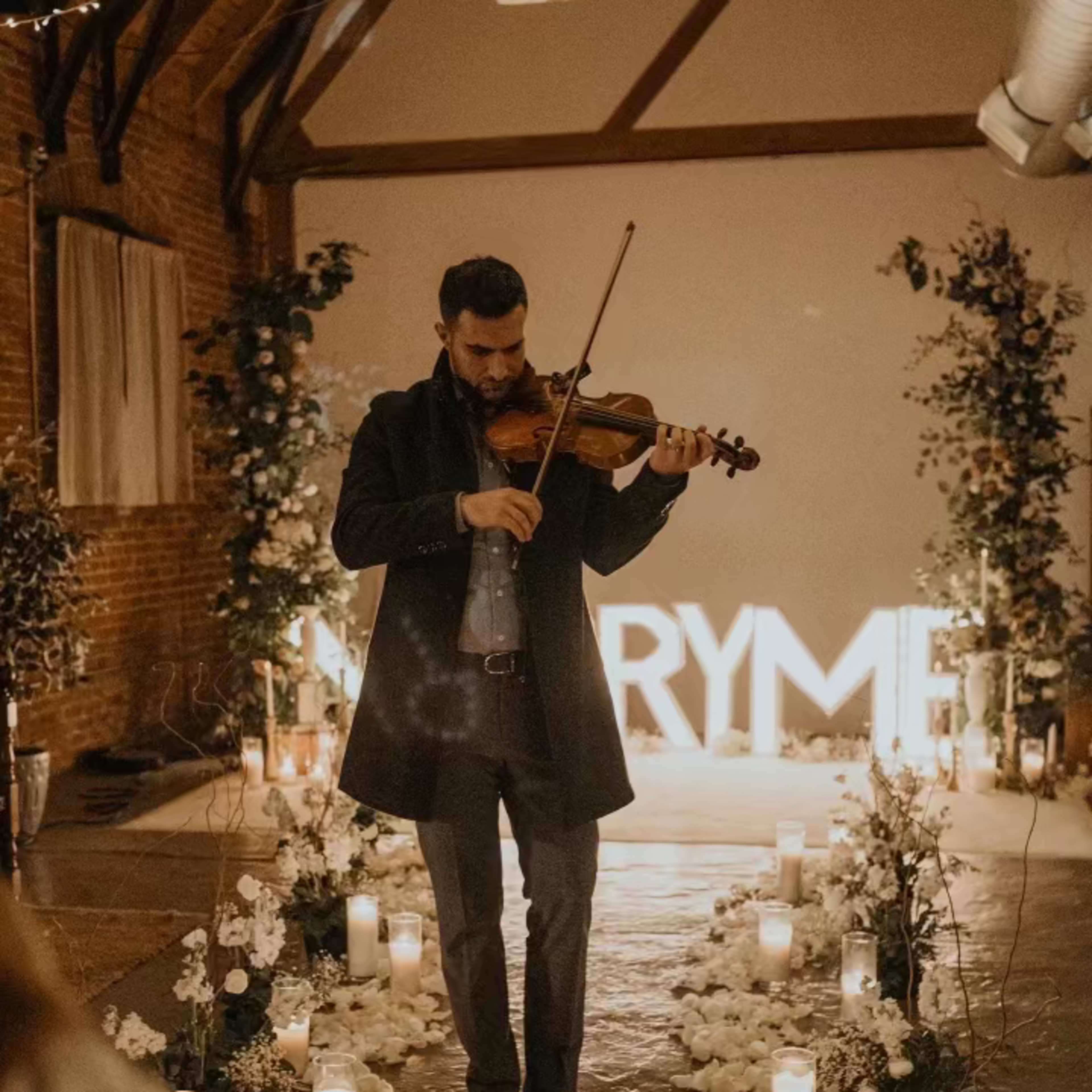 A man in a black coat plays the violin in a venue adorned with candles and flowers, with a large illuminated sign in the background.