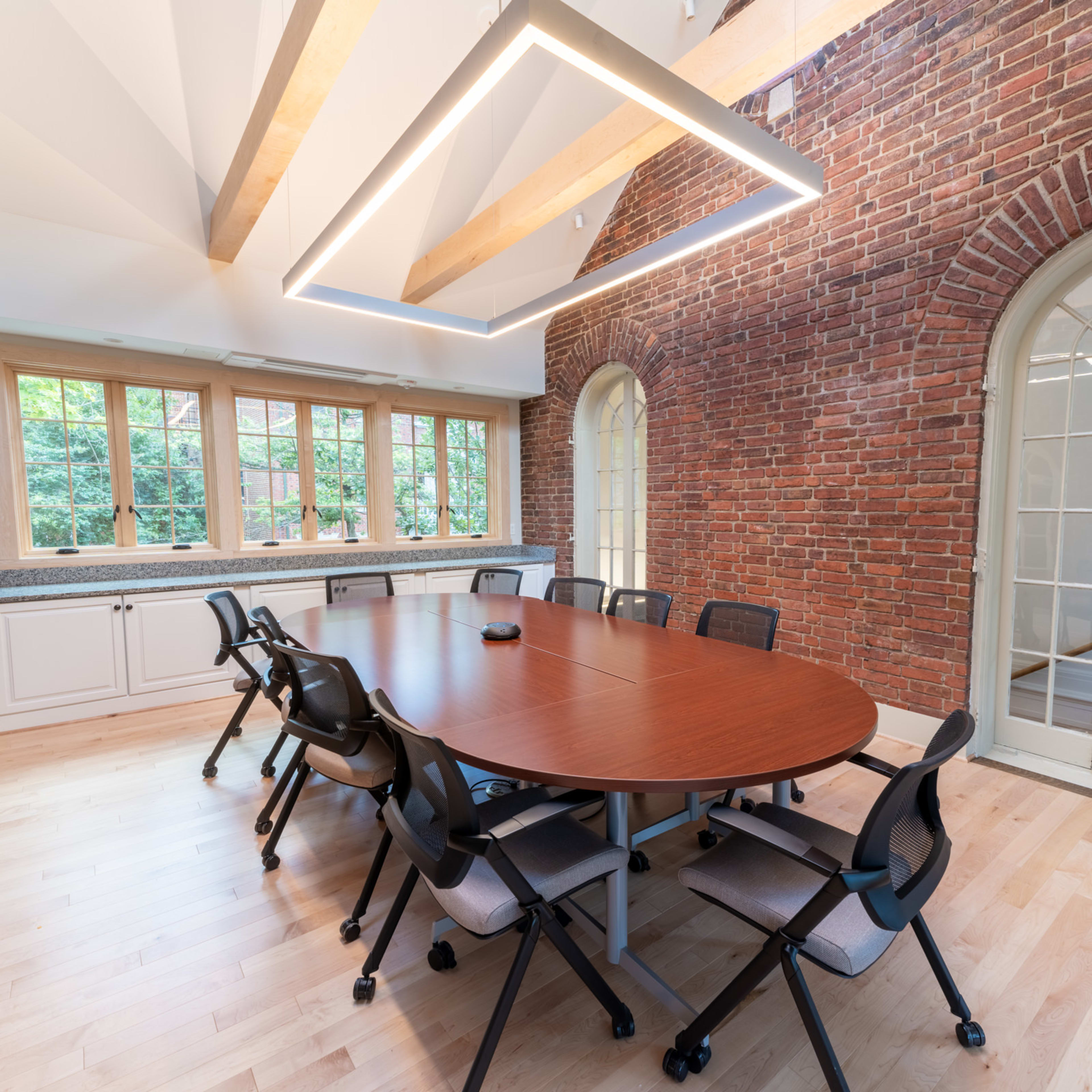A modern conference room featuring a round wooden table surrounded by black chairs, with exposed brick walls and large windows allowing natural light.