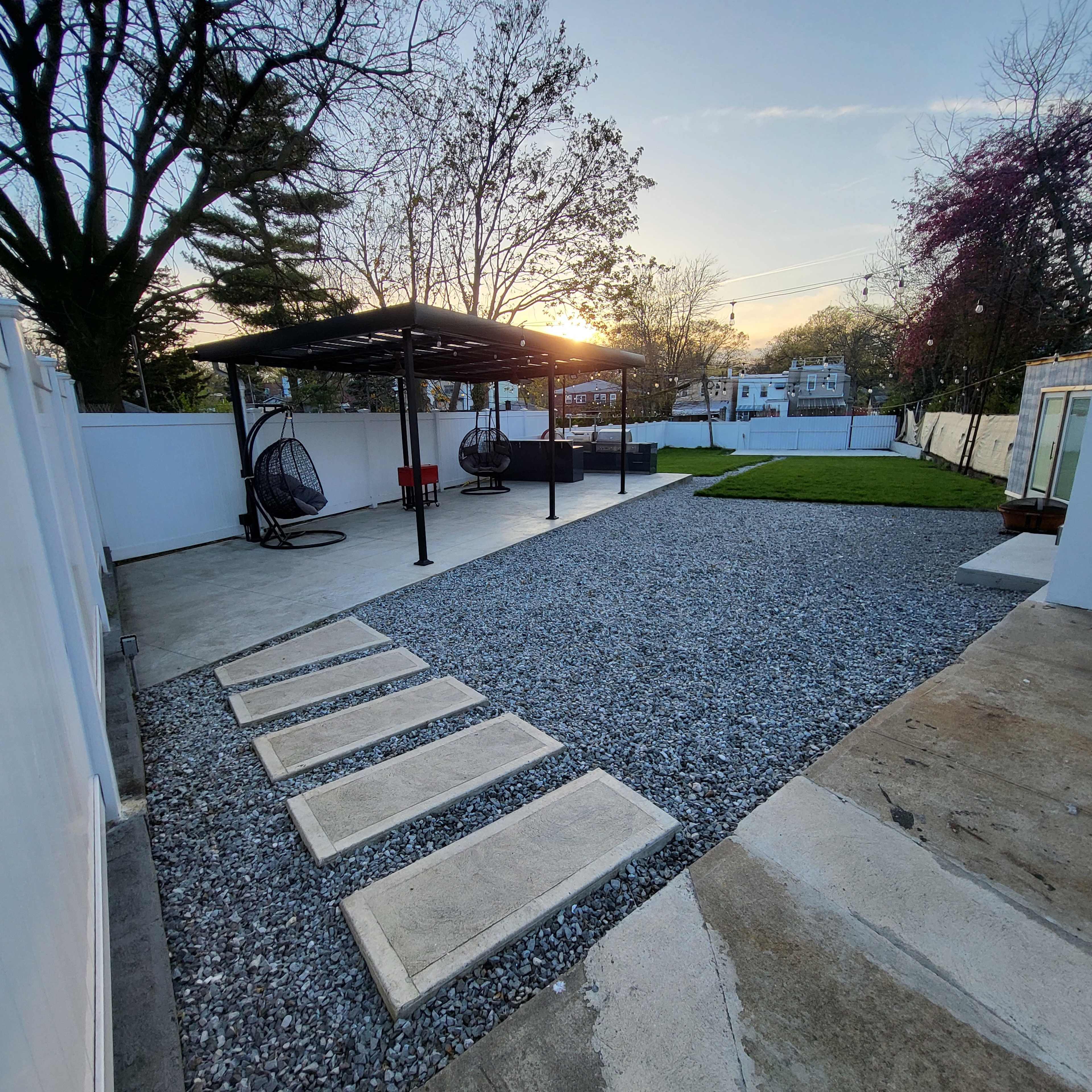 A backyard with a gravel area, concrete stepping stones, a covered seating area, and a green lawn under a sunset sky.