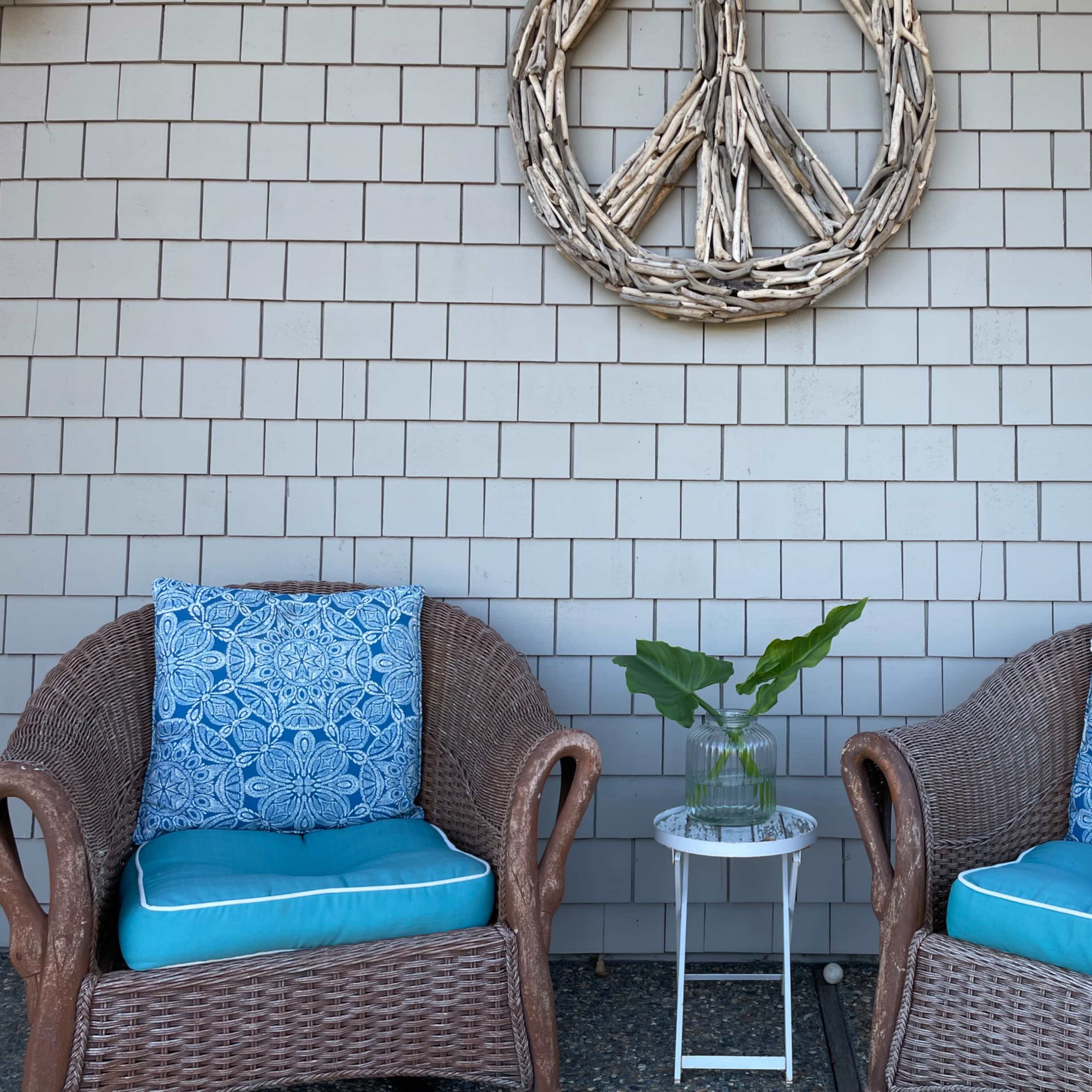 Two wicker chairs with blue cushions sit beside a small round table, featuring a vase with a plant, against a shingle-style wall adorned with a driftwood peace sign.