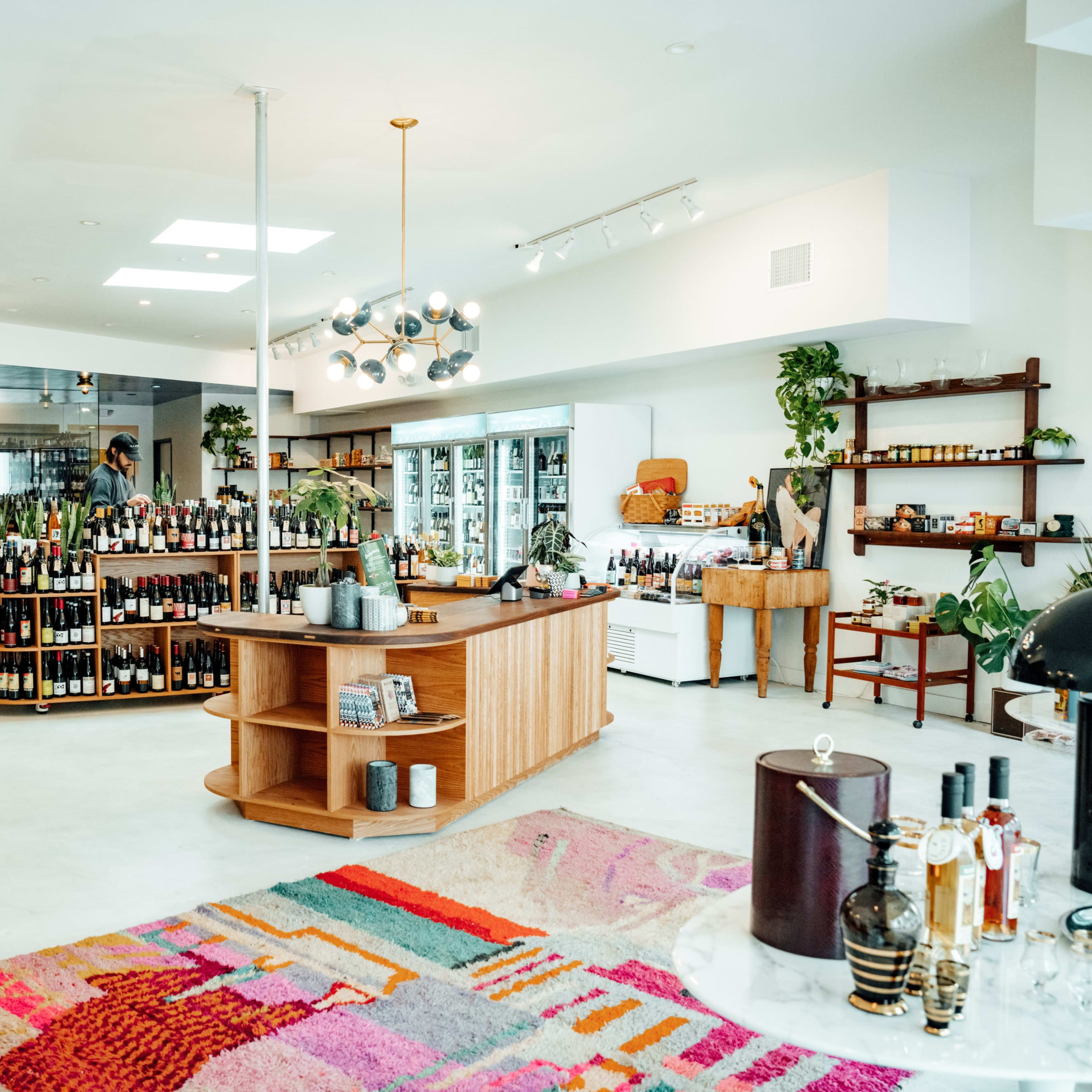 A wine shop interior featuring wooden shelves stocked with various bottles, a central display table, and potted plants throughout the space.