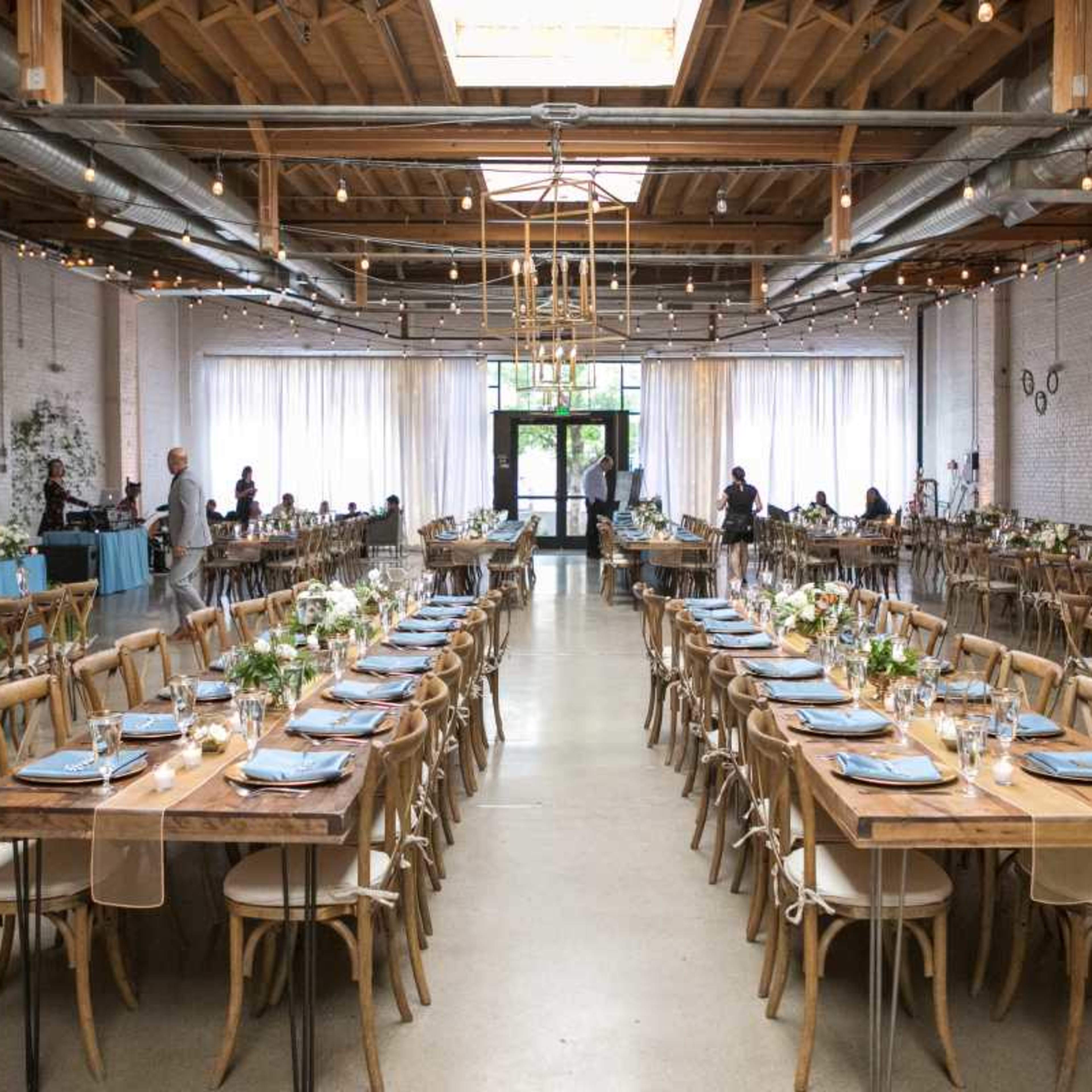 A spacious banquet hall is set up with long wooden tables adorned with floral centerpieces and blue table settings, illuminated by overhead lights and large windows.
