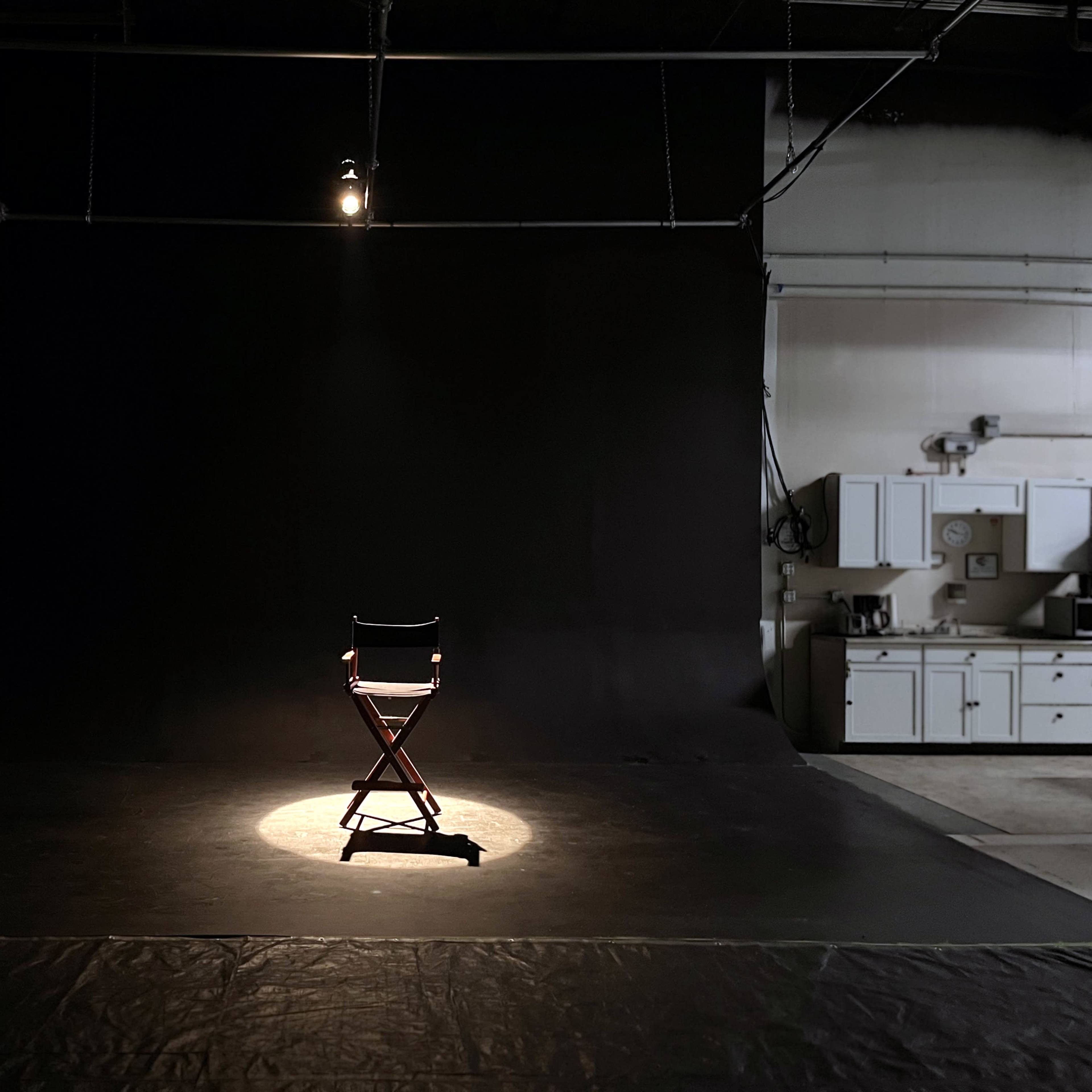 A single director's chair is illuminated by a spotlight on a dark stage with a black backdrop.
