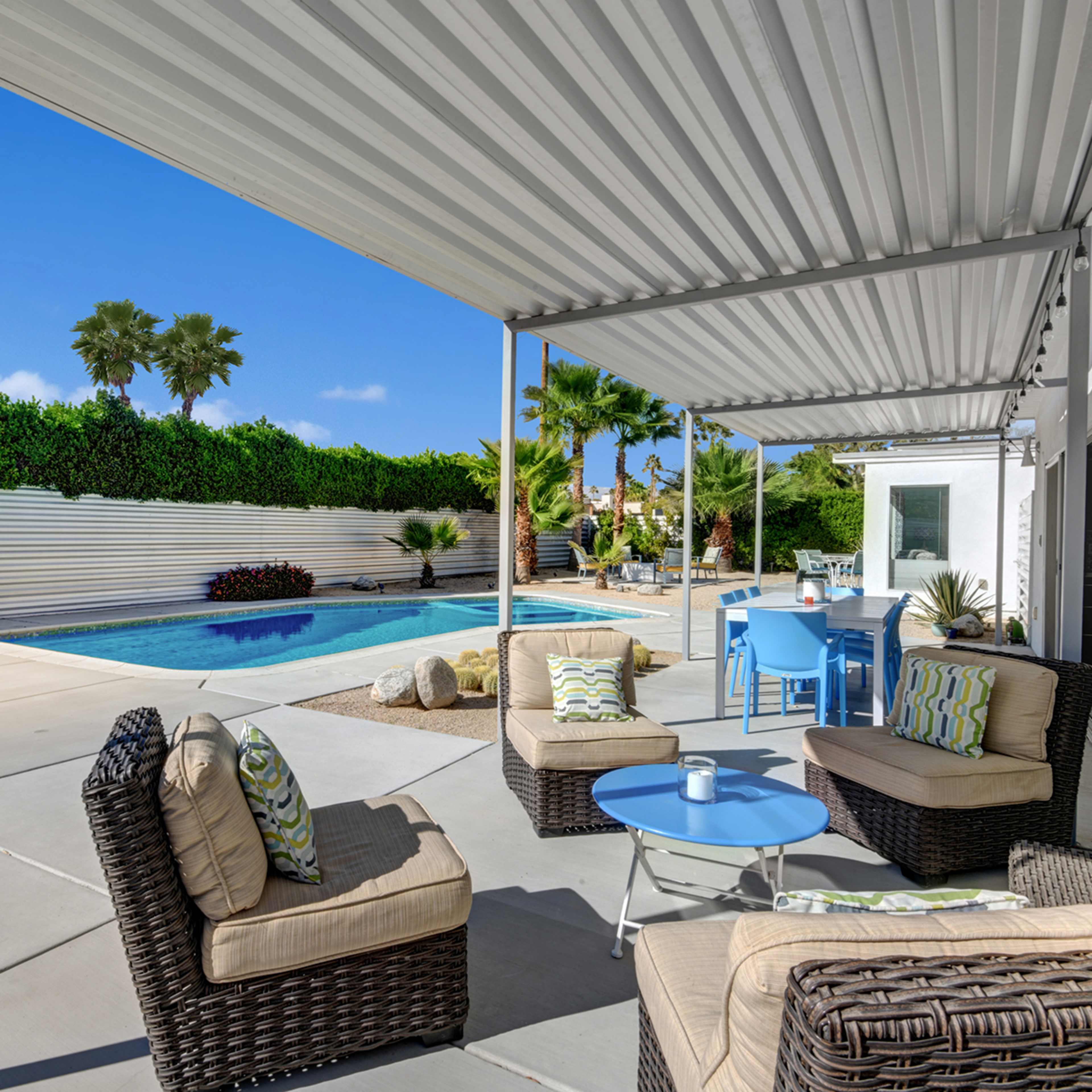 A patio features comfortable seating arrangements and a swimming pool, surrounded by greenery and a bright sky.