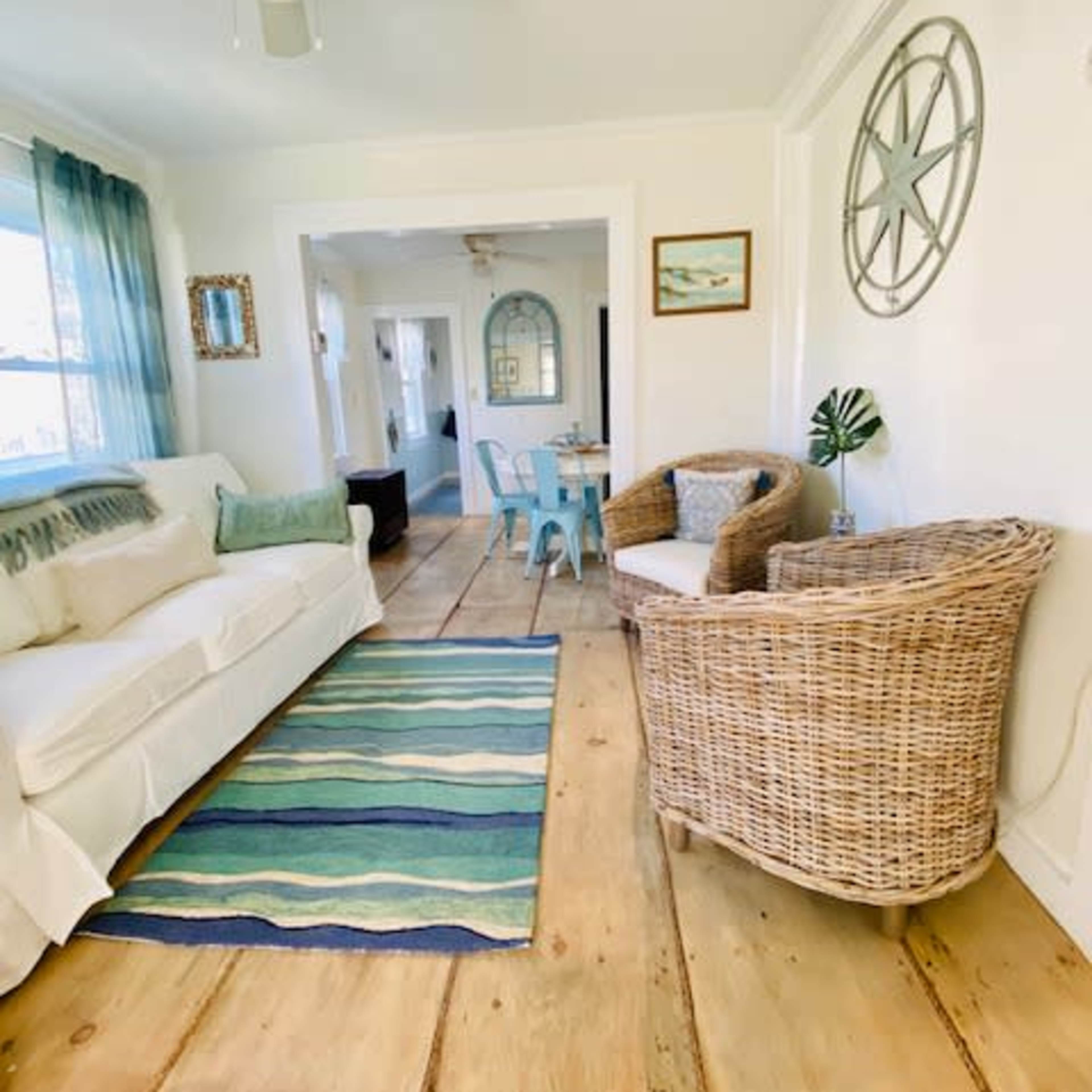 A light-filled living room with a white sofa, a striped rug, and wicker furniture, leading to a dining area in the background.