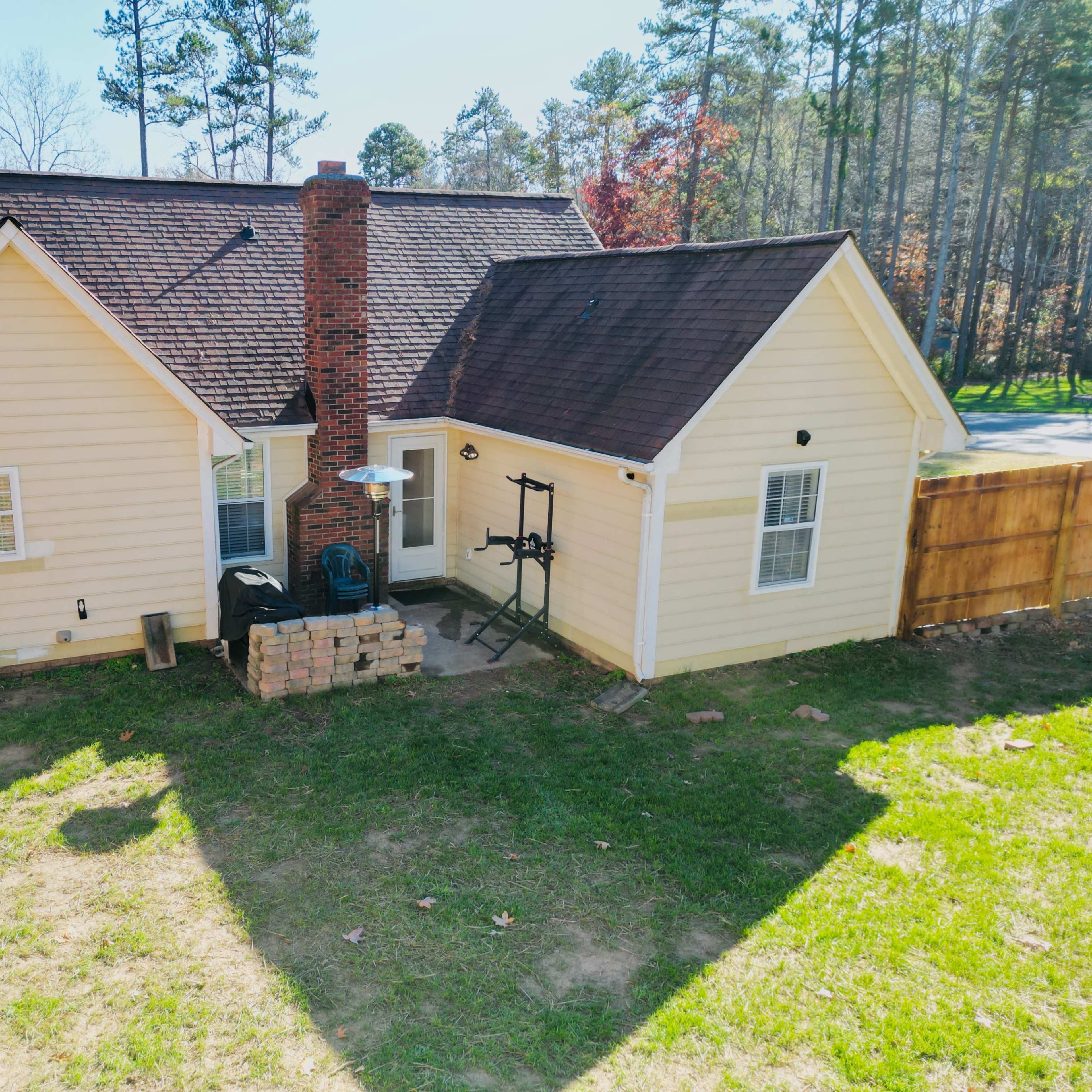 A yellow single-story house with a brick chimney, surrounded by trees, and a small patio area with a grill.