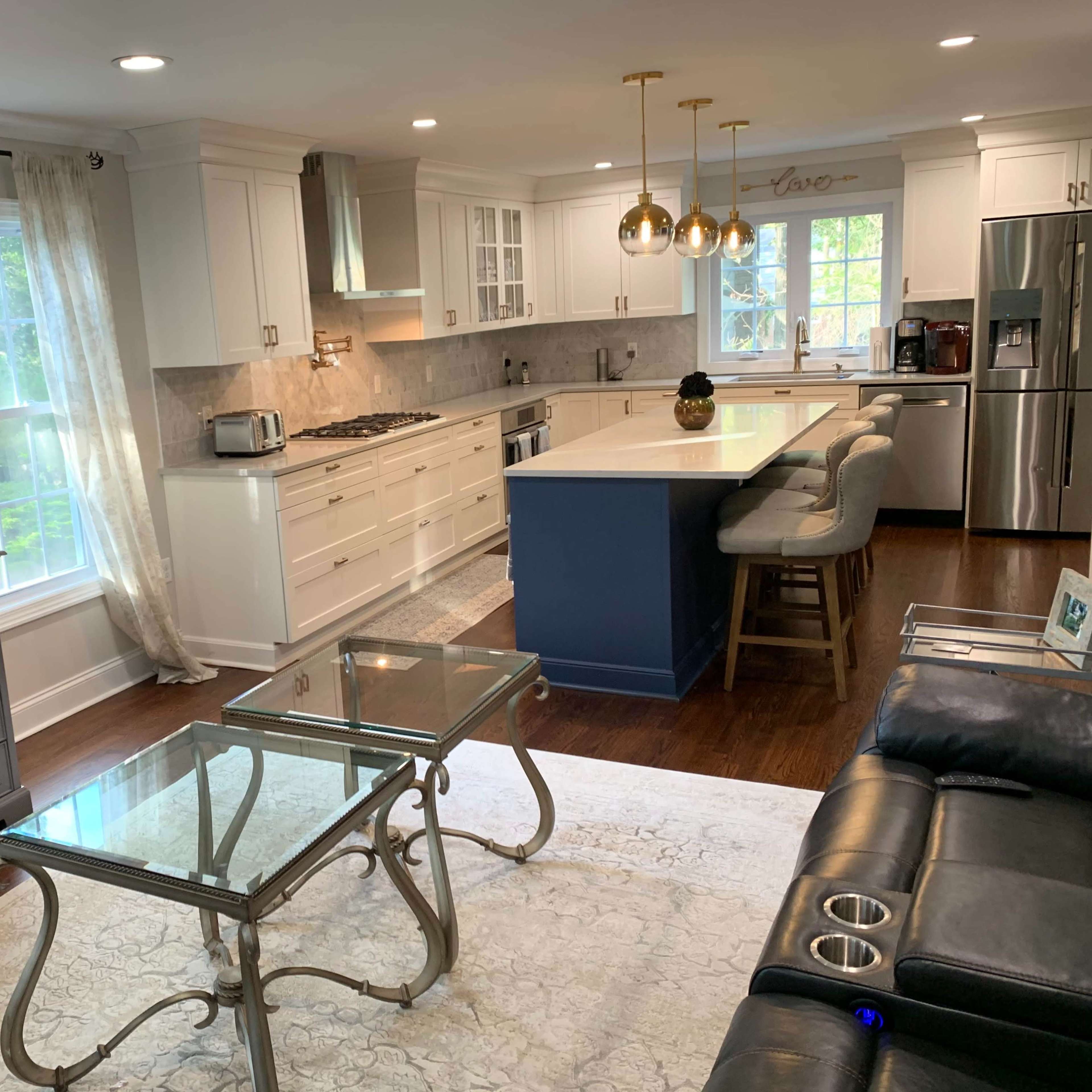 A modern kitchen and living area featuring white cabinetry, stainless steel appliances, a large island with seating, and a glass coffee table on a patterned rug.