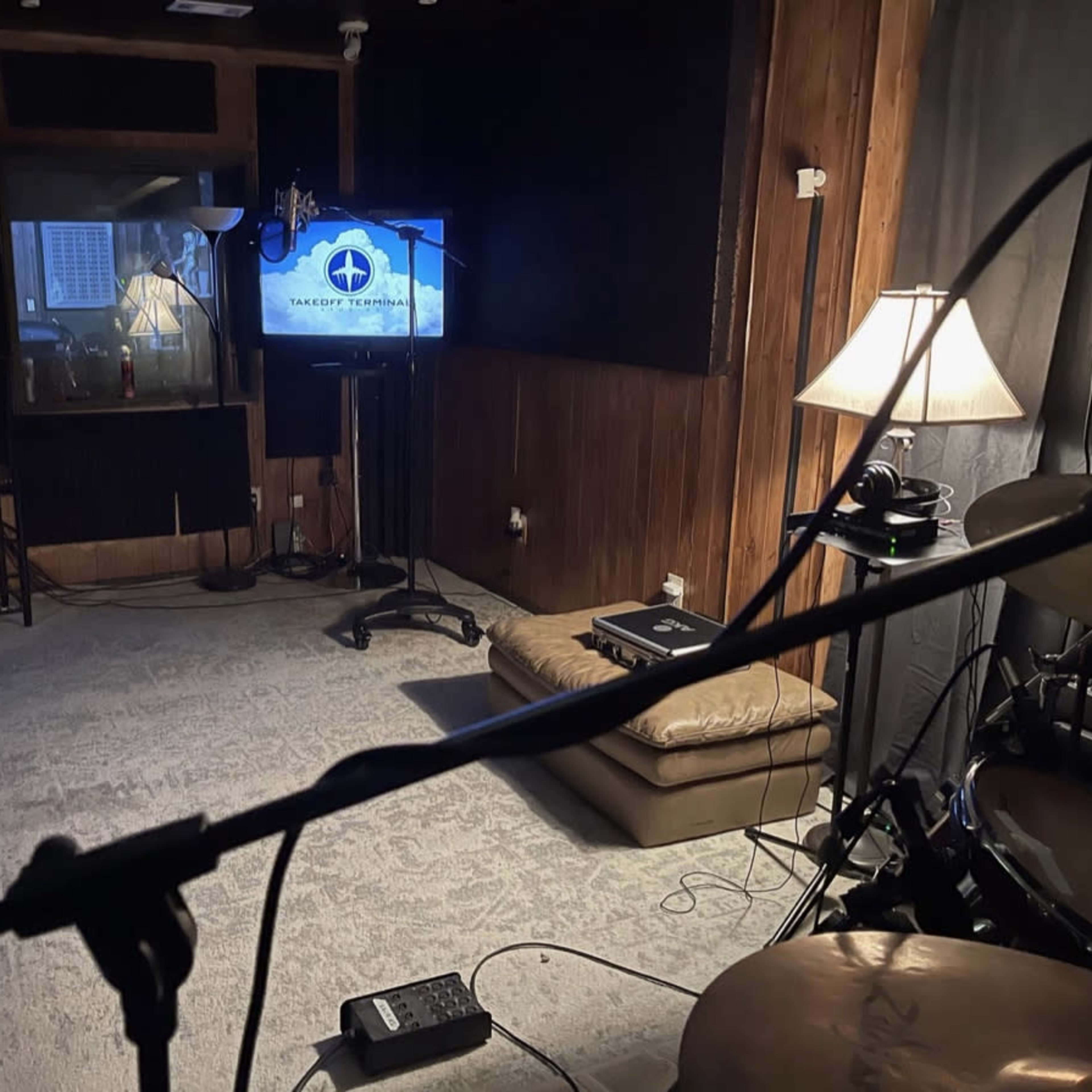 A music studio features a couch, TV, and lamp, with a drum set and microphone in the foreground.