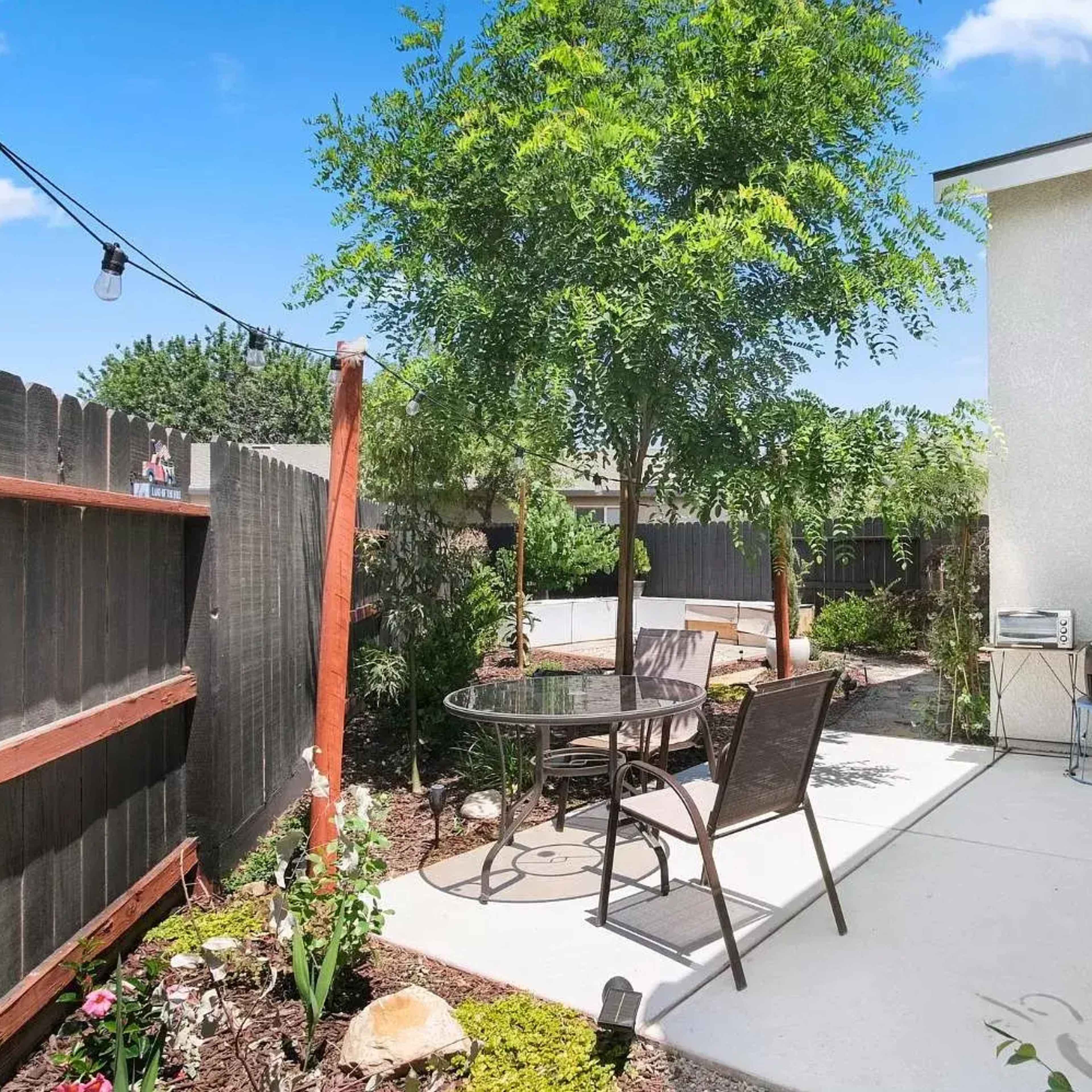 A patio area features a table and chairs surrounded by greenery and a wooden fence.