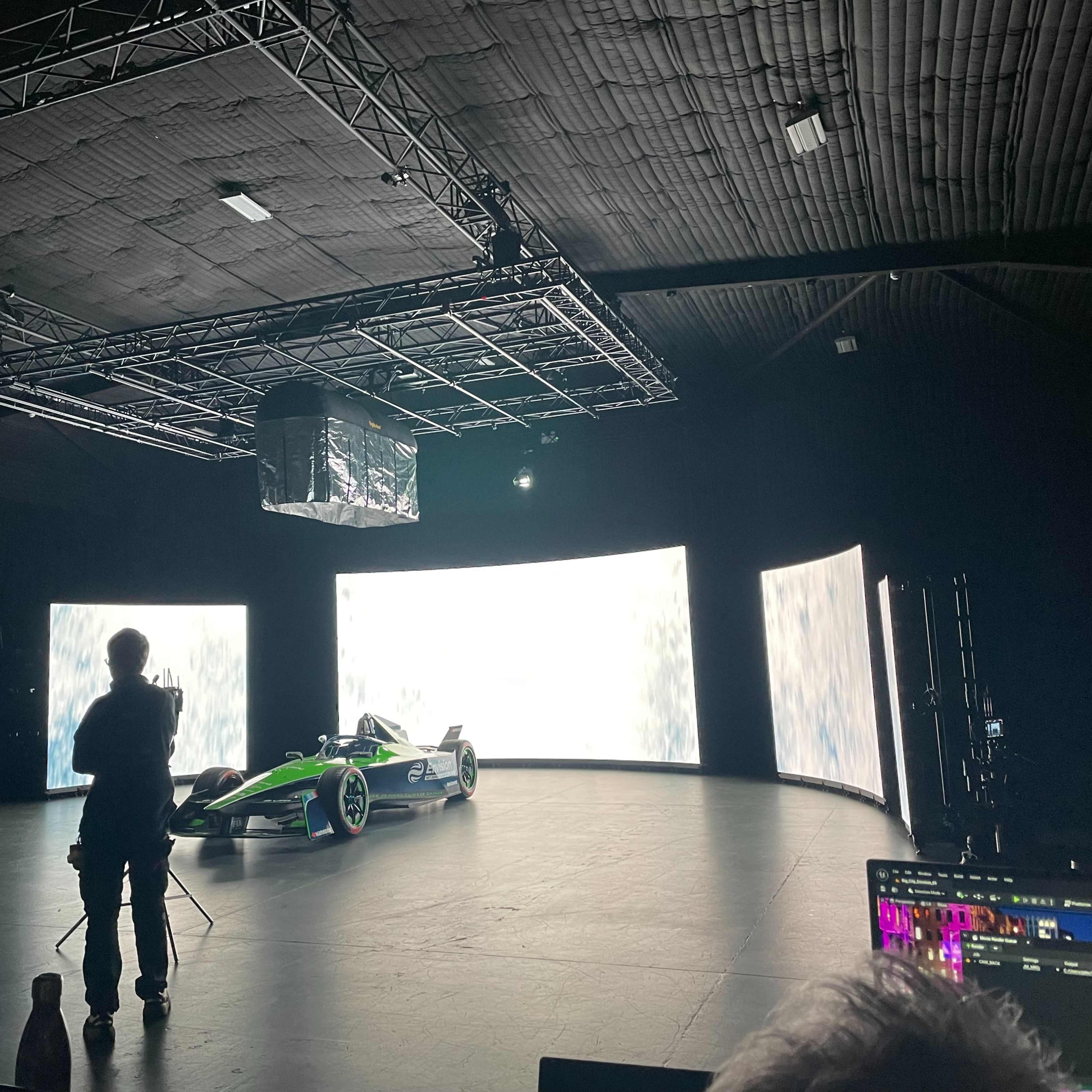 A race car is positioned in a studio with large screens displaying dynamic visual effects while a person stands nearby.