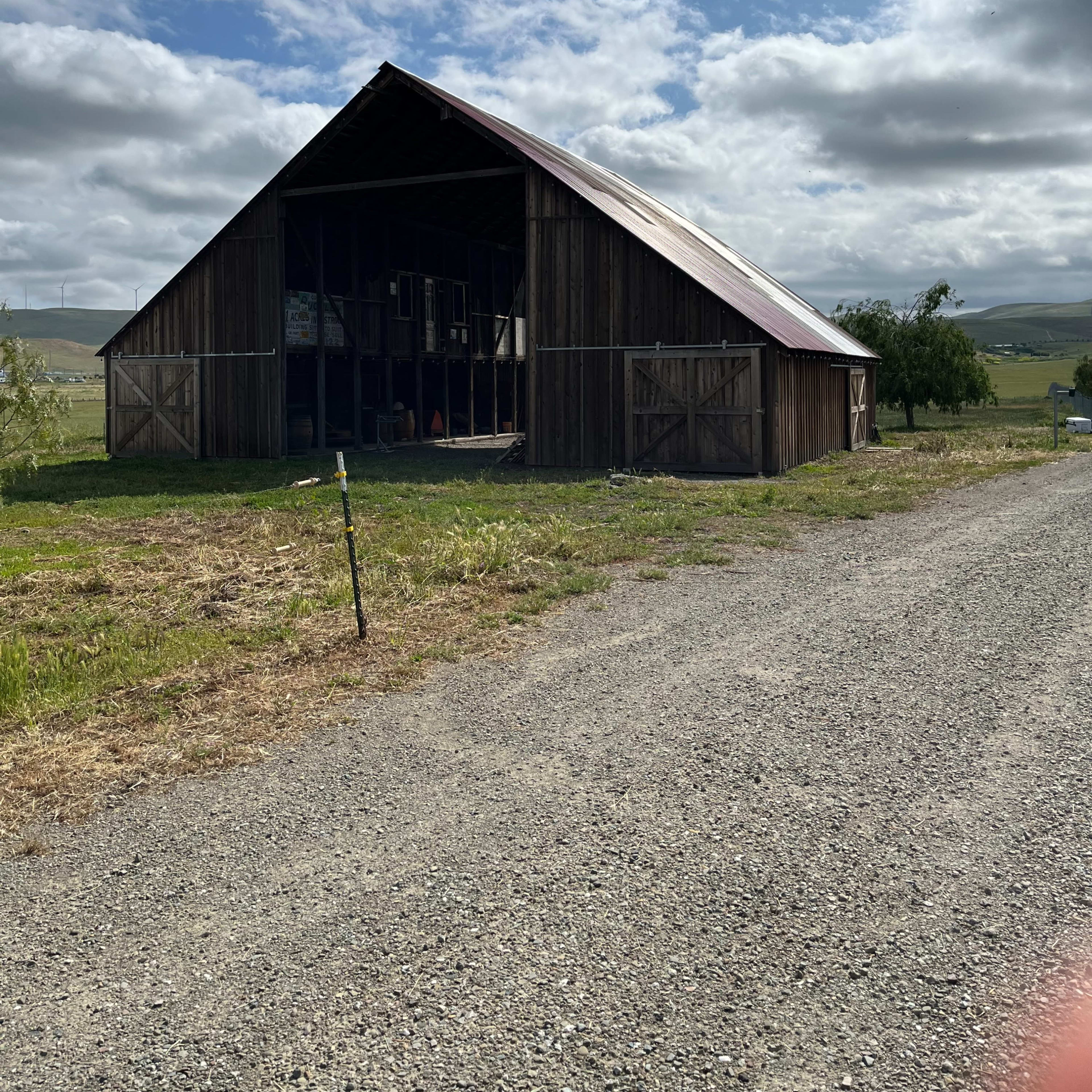 A wooden barn with a metal roof is situated next to a gravel road under a cloudy sky.
