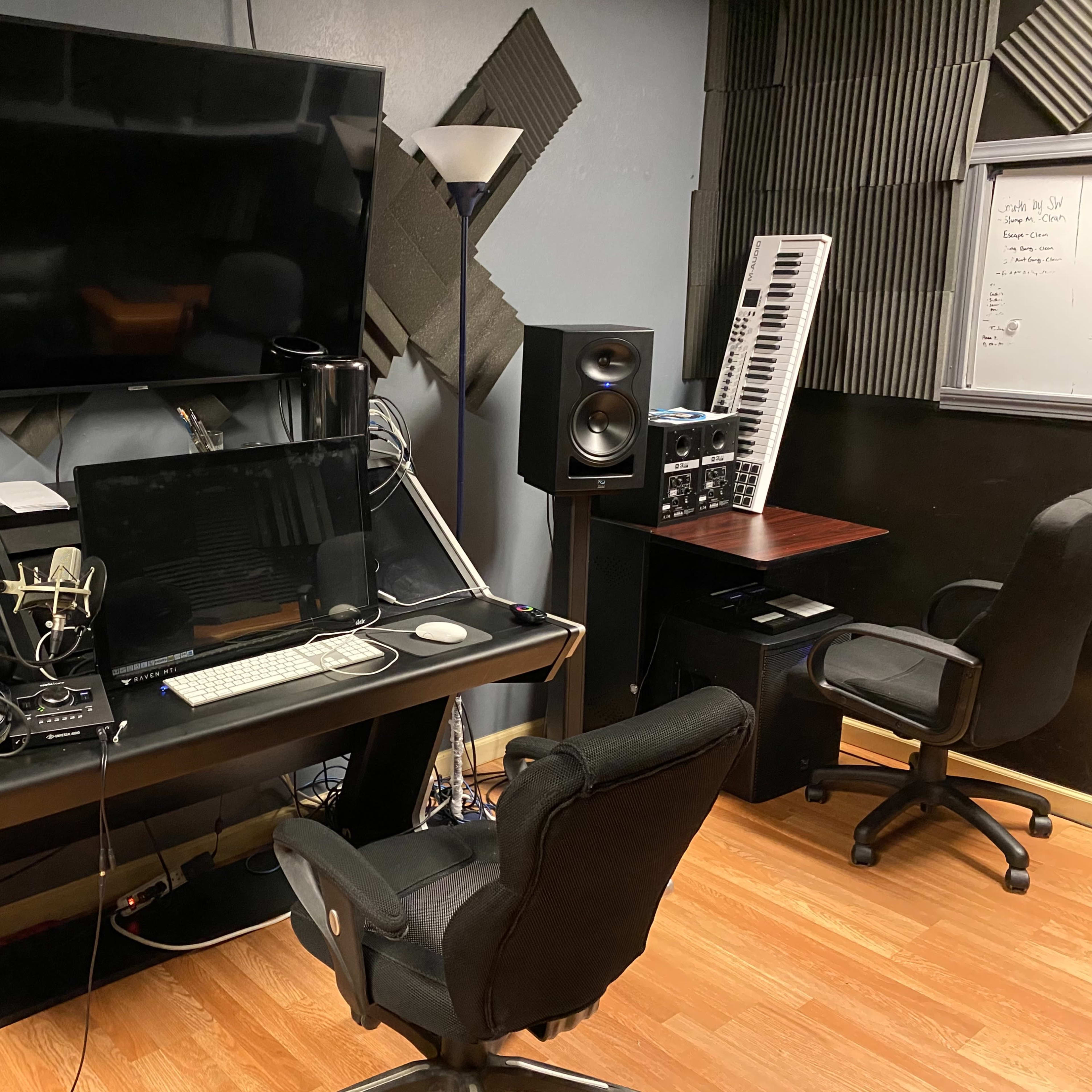 A music production studio featuring a desk with a computer, speakers, and a microphone, along with a keyboard and additional equipment on the walls.