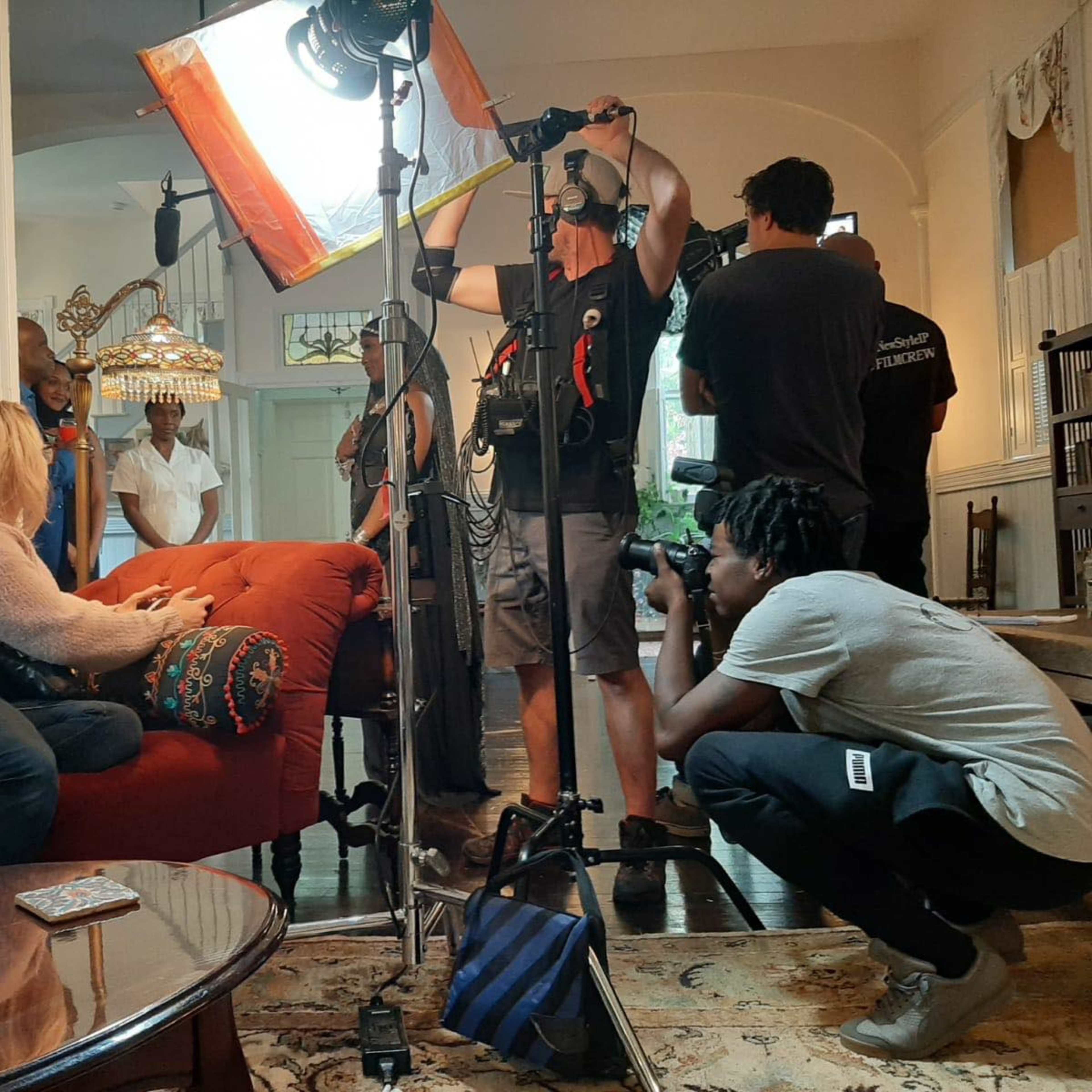 A film crew sets up equipment in a bright living room while actors prepare for a scene.