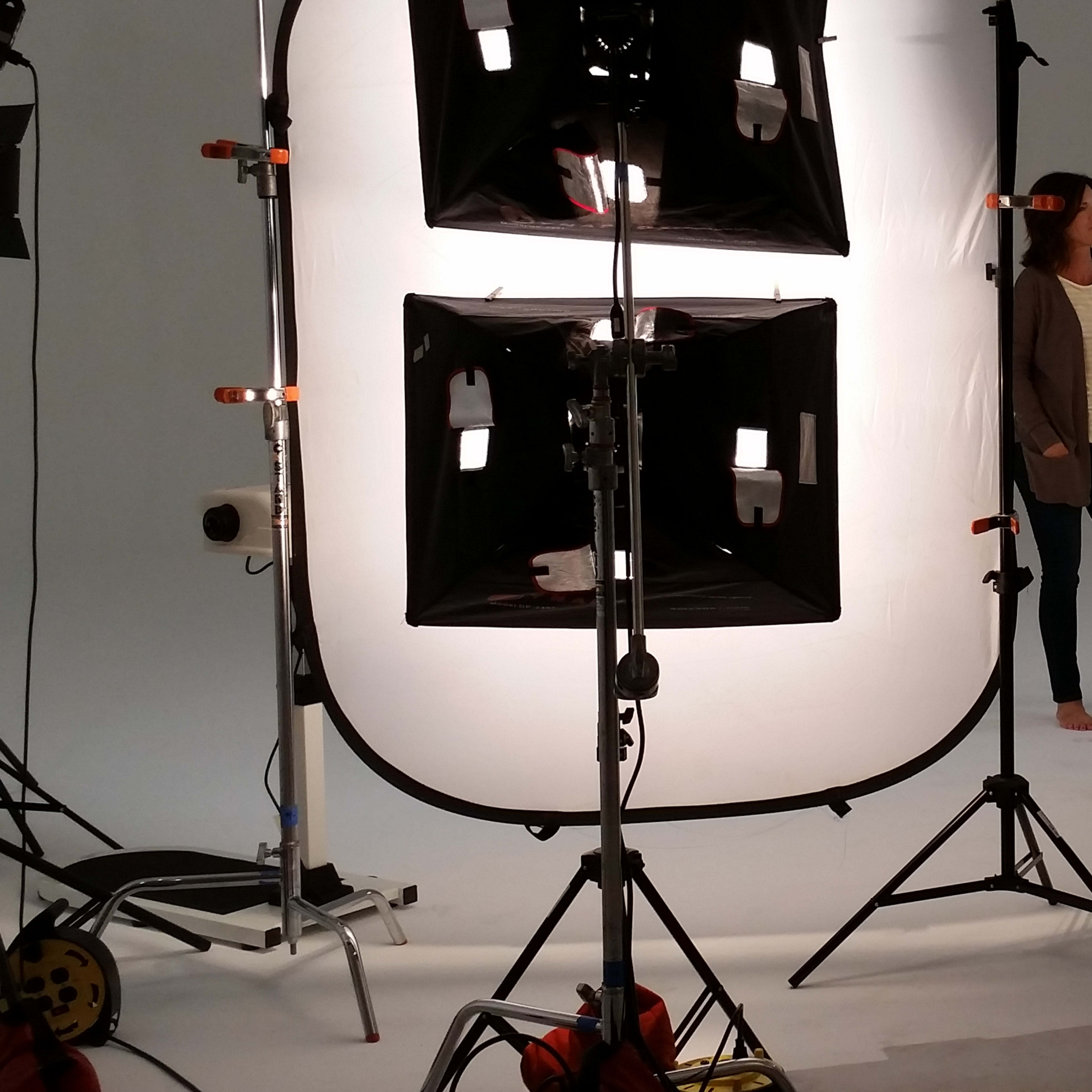 A model stands barefoot in a well-lit photography studio surrounded by multiple lights and cameras.