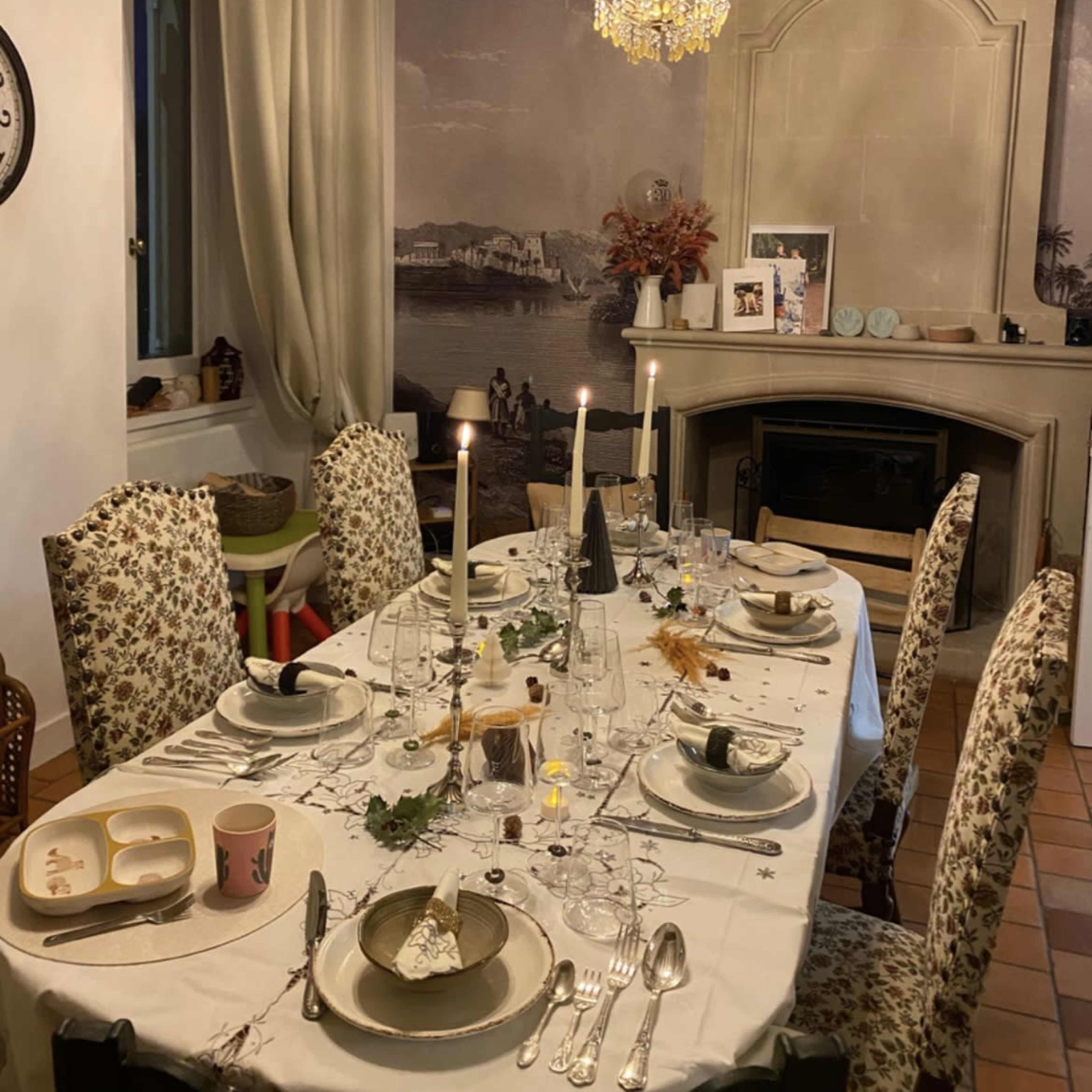 A dining table is set with multiple place settings, candles, and decorative elements in a warmly lit room featuring wooden beams and a fireplace.