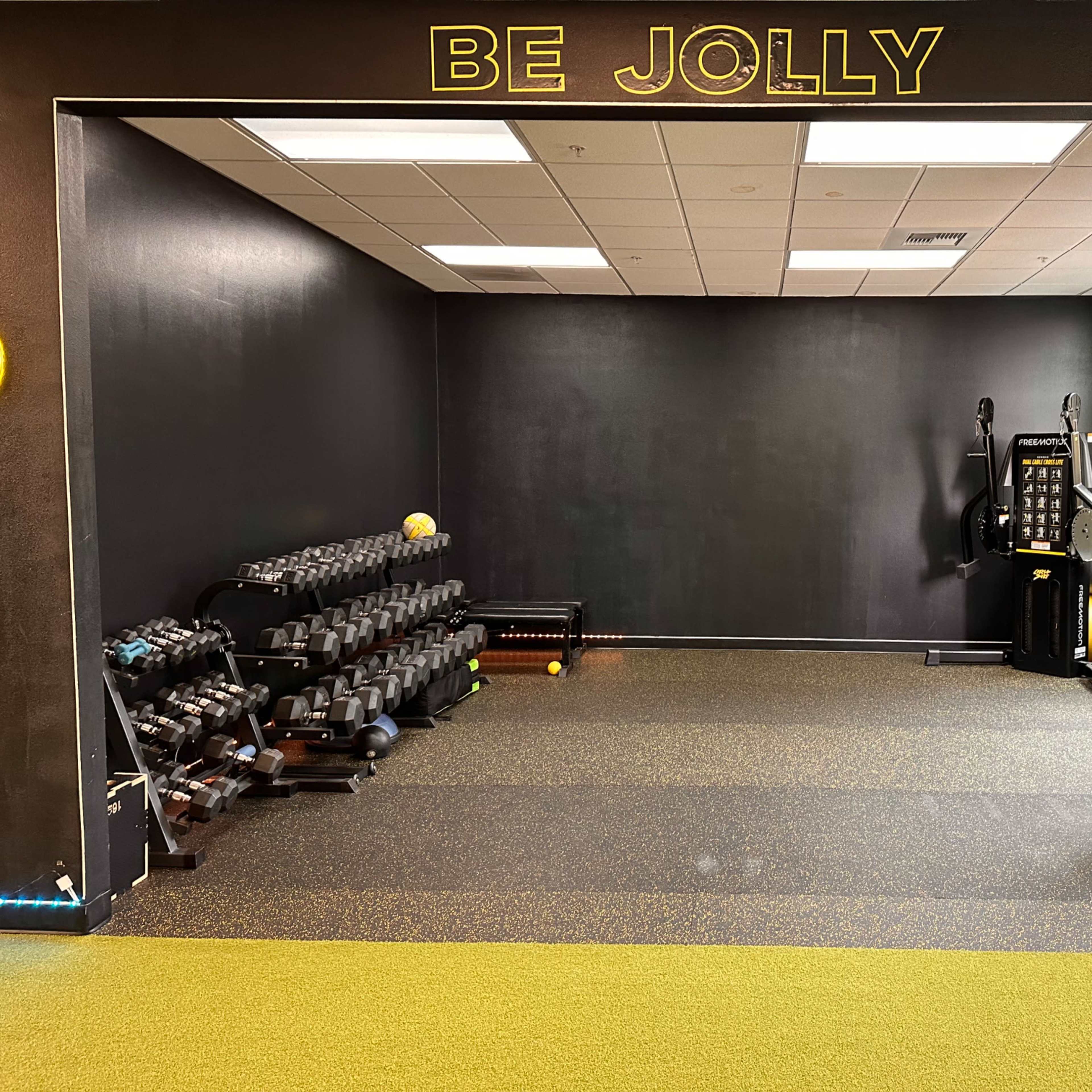 A fitness area with weights on racks, a stationary bike, and exercise equipment against a dark wall featuring the words "BE JOLLY."