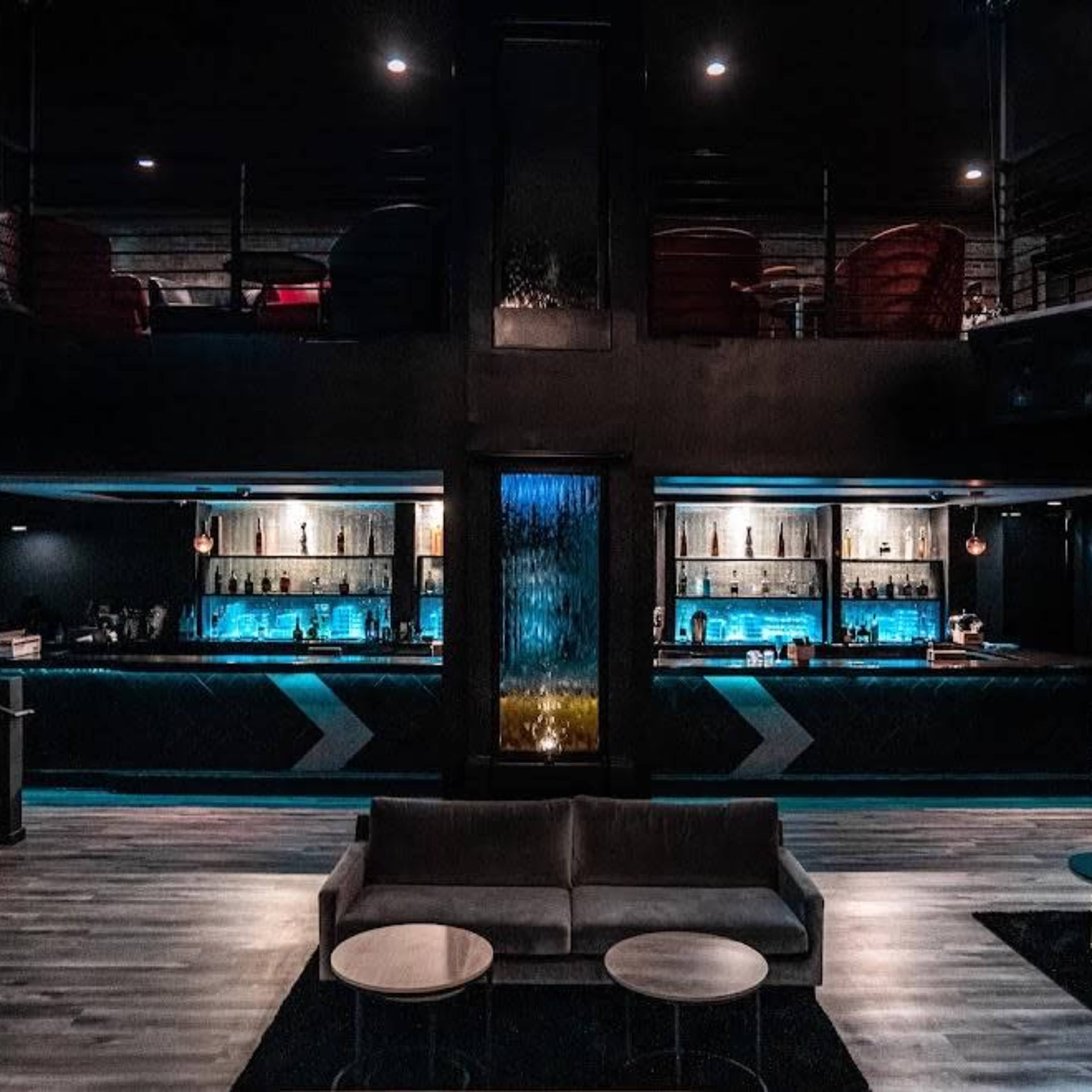 A dimly lit bar area with a waterfall feature, a long illuminated bar, and seating arranged on two levels.