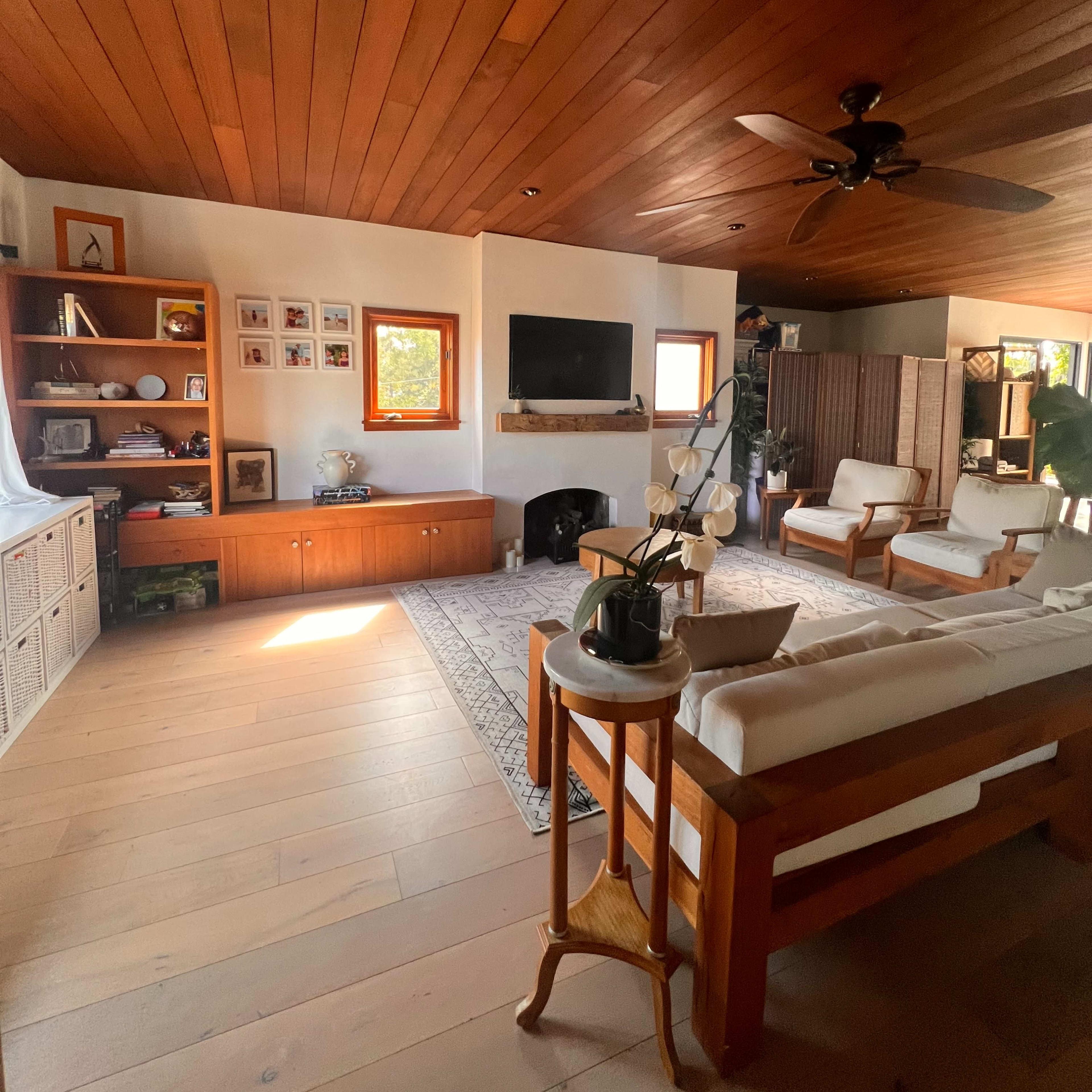 A bright living room featuring wooden walls and ceiling, a seating area with two chairs and a couch, a television mounted on the wall, and a decorative bookshelf.