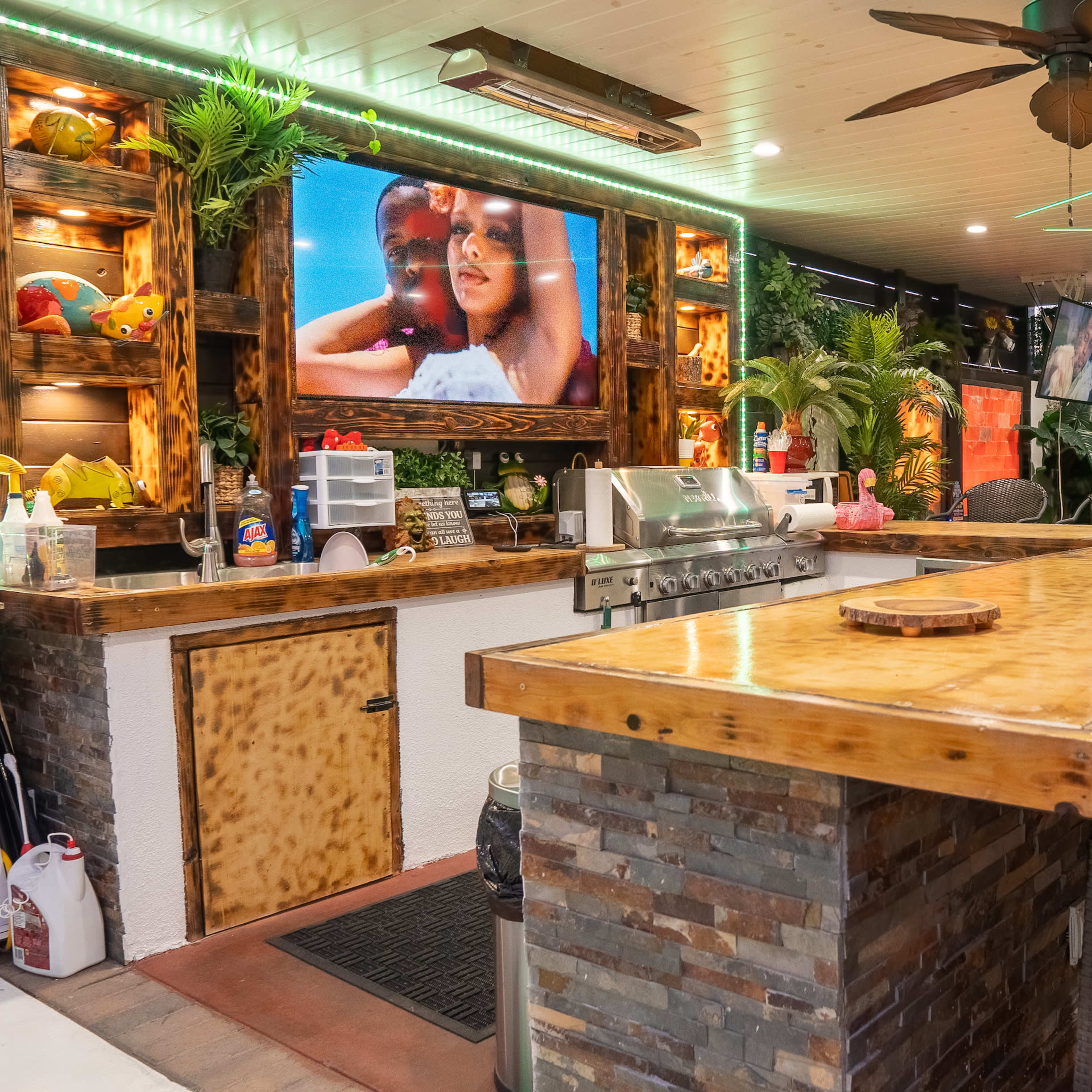 A well-decorated outdoor bar area featuring a wooden countertop, comfortable seating, and a large TV mounted on the wall.