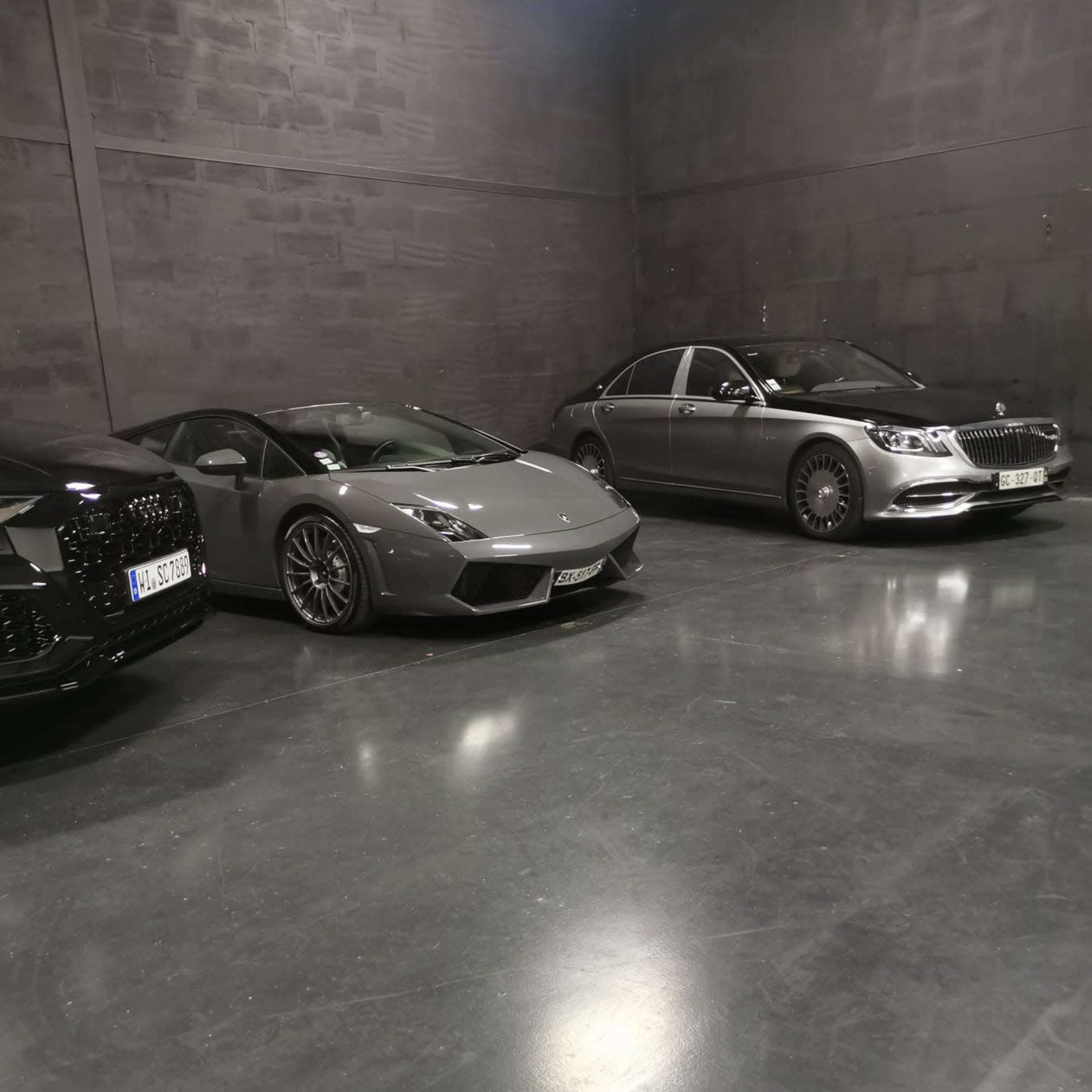 Three luxury cars parked in a dimly lit garage, including a gray Lamborghini in the center, alongside a sleek black car on the left and a silver Mercedes-Benz on the right.
