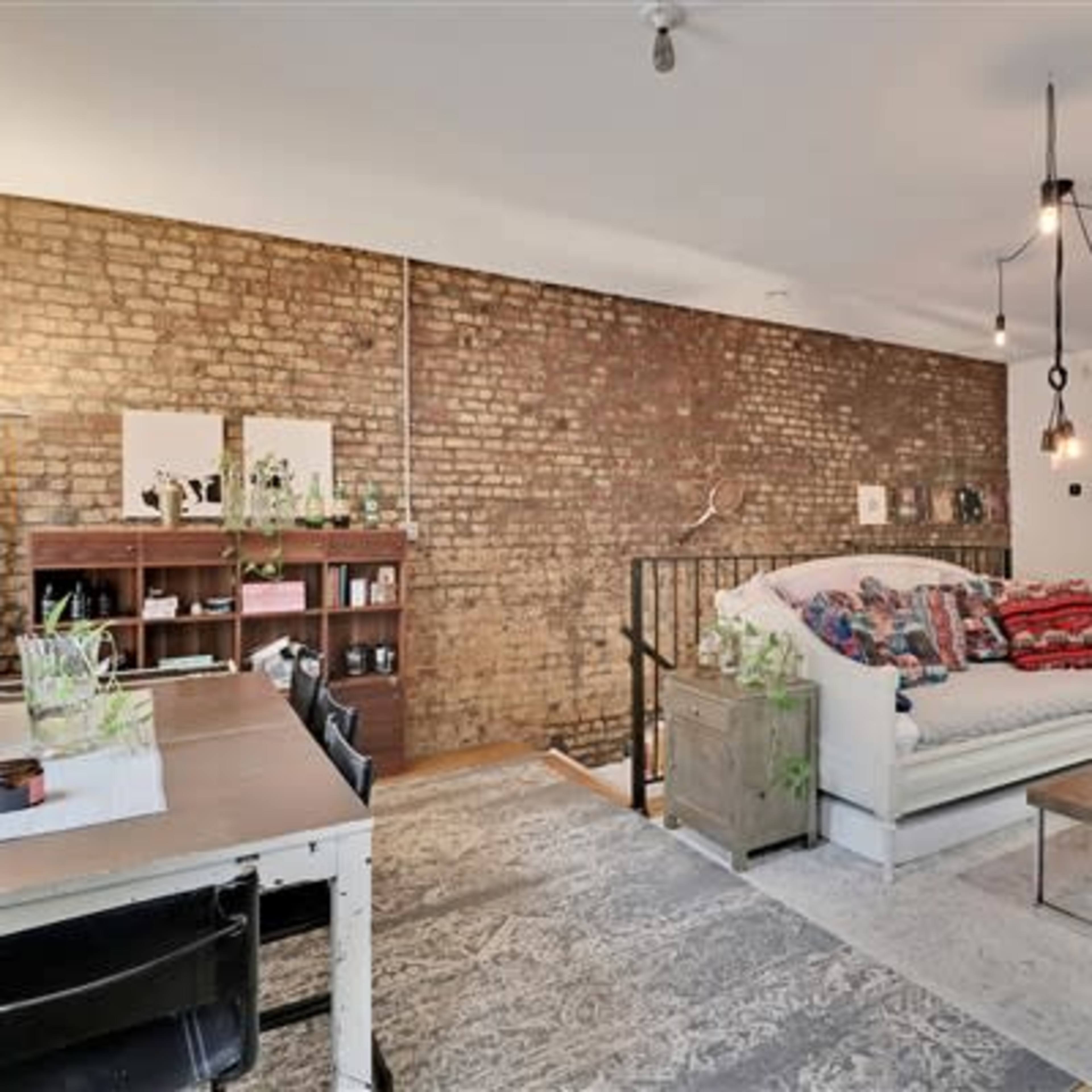 A living area with a brick wall, a white sofa adorned with colorful throw blankets, a wooden bookshelf, and a coffee table on a patterned rug.