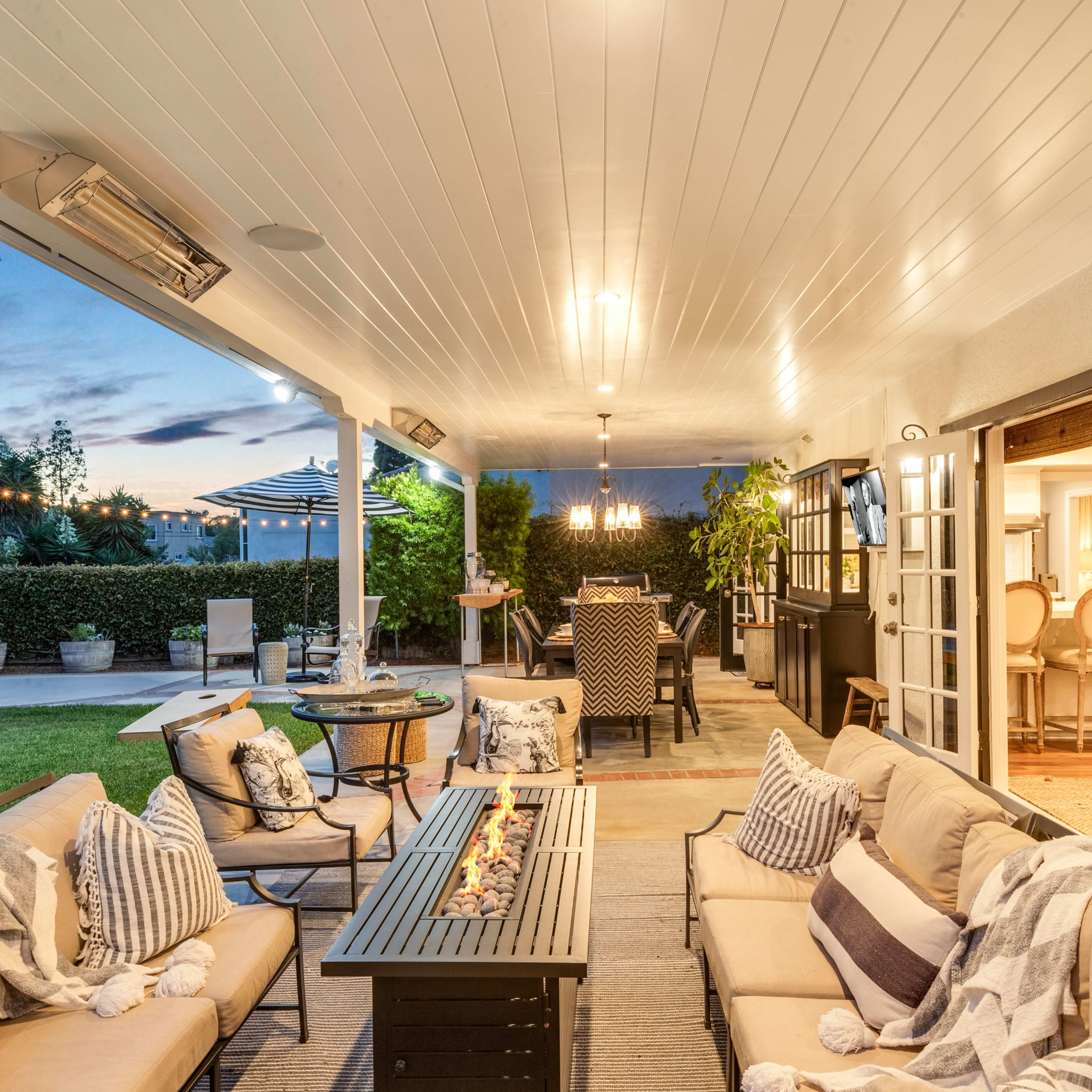 A covered outdoor patio featuring a seating area with a fire table, potted plants, and a dining space visible in the background.