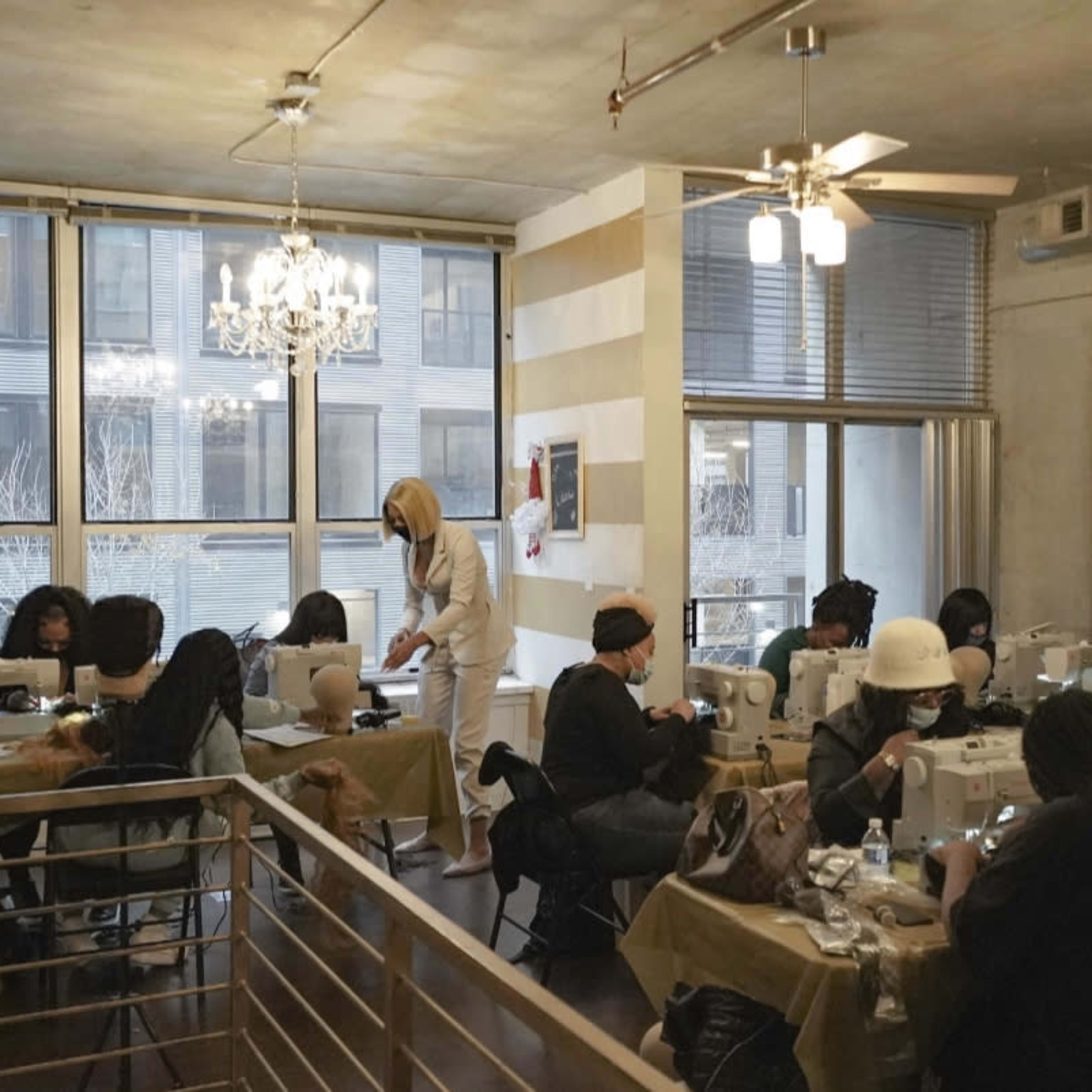 A group of people is engaged in a sewing workshop, using sewing machines at tables in a well-lit room with large windows.