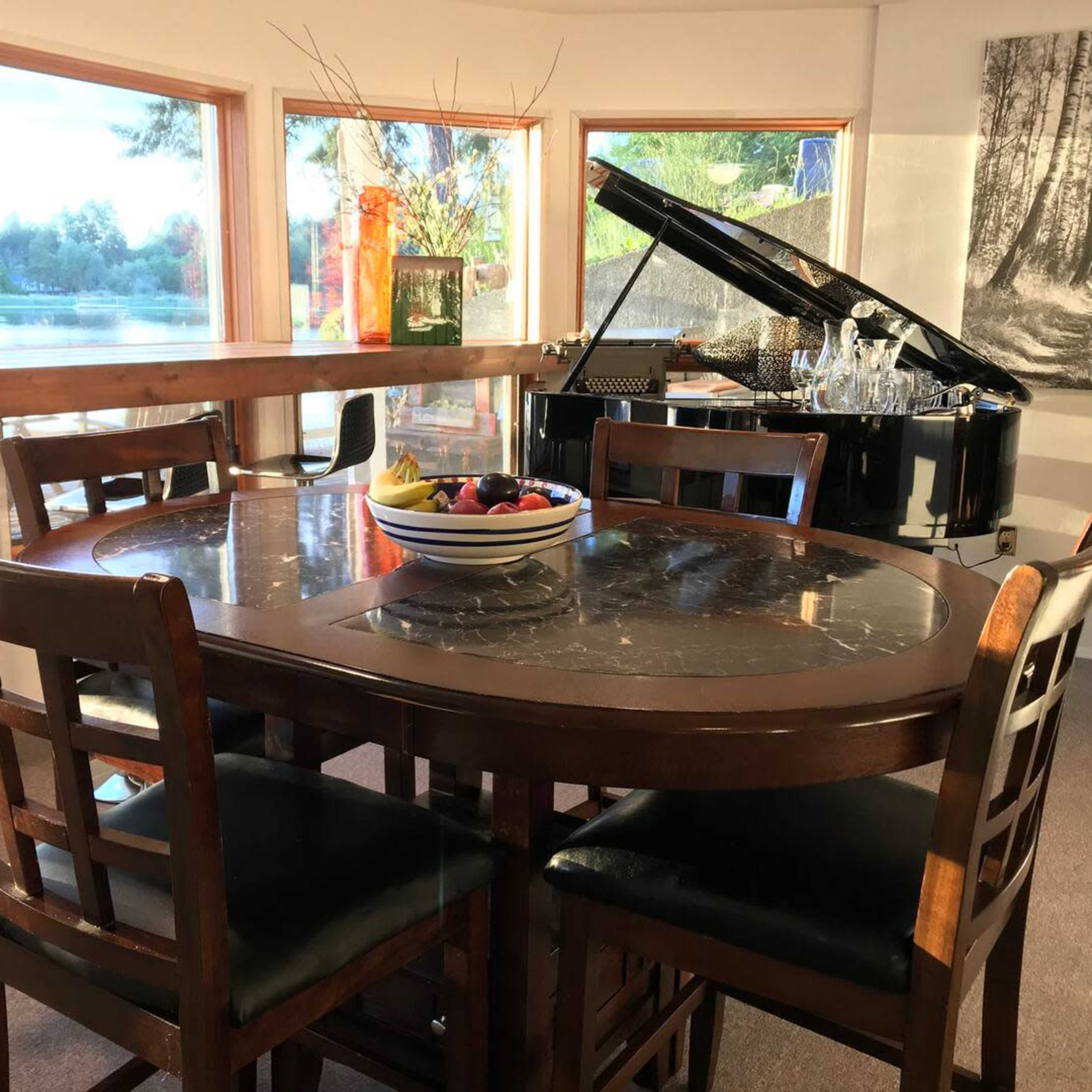 A round wooden dining table with four chairs is positioned near a large window overlooking a scenic view, beside a black grand piano and a display of glassware.