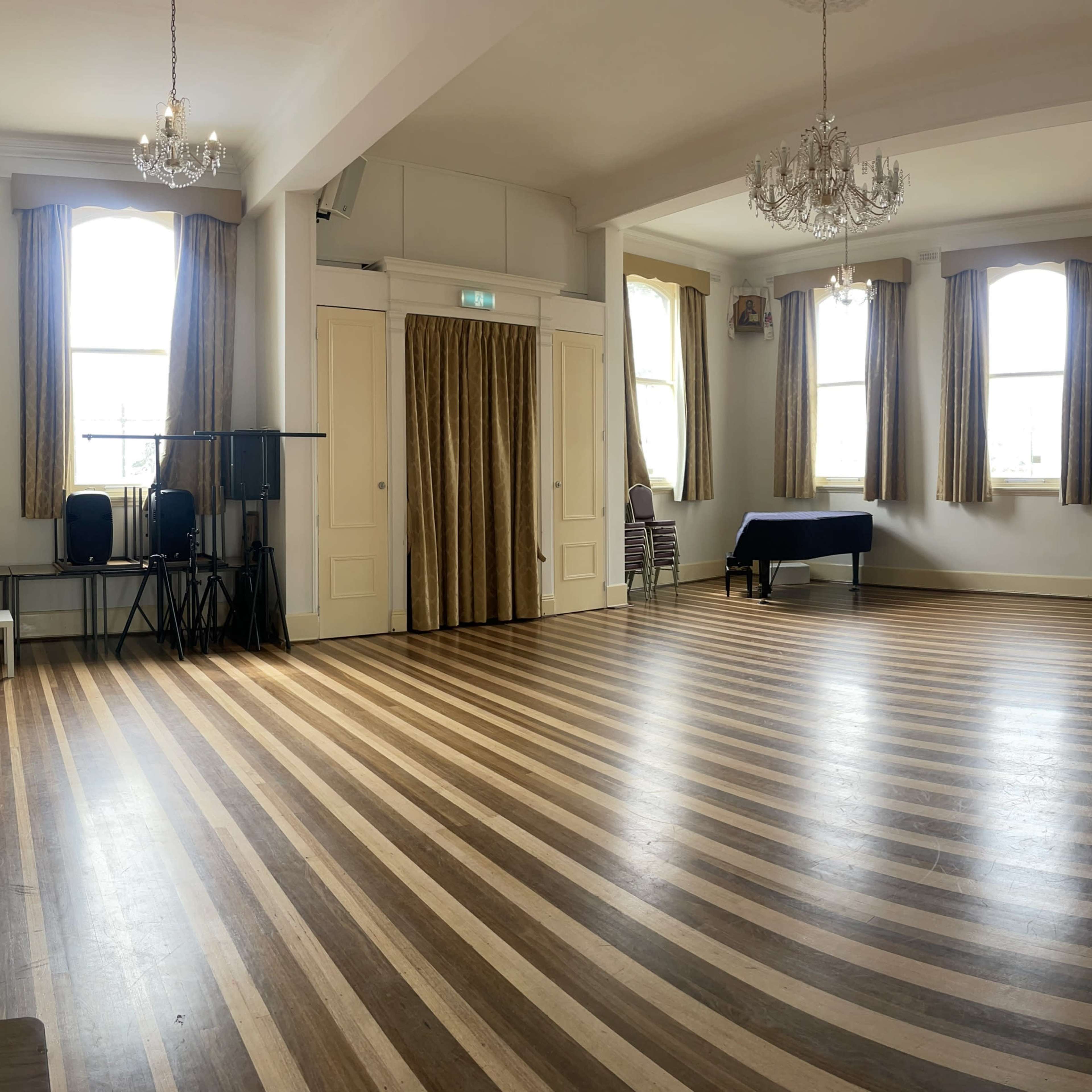 A spacious, empty room with polished wooden floors and large windows, featuring cream-colored walls and two chandeliers.